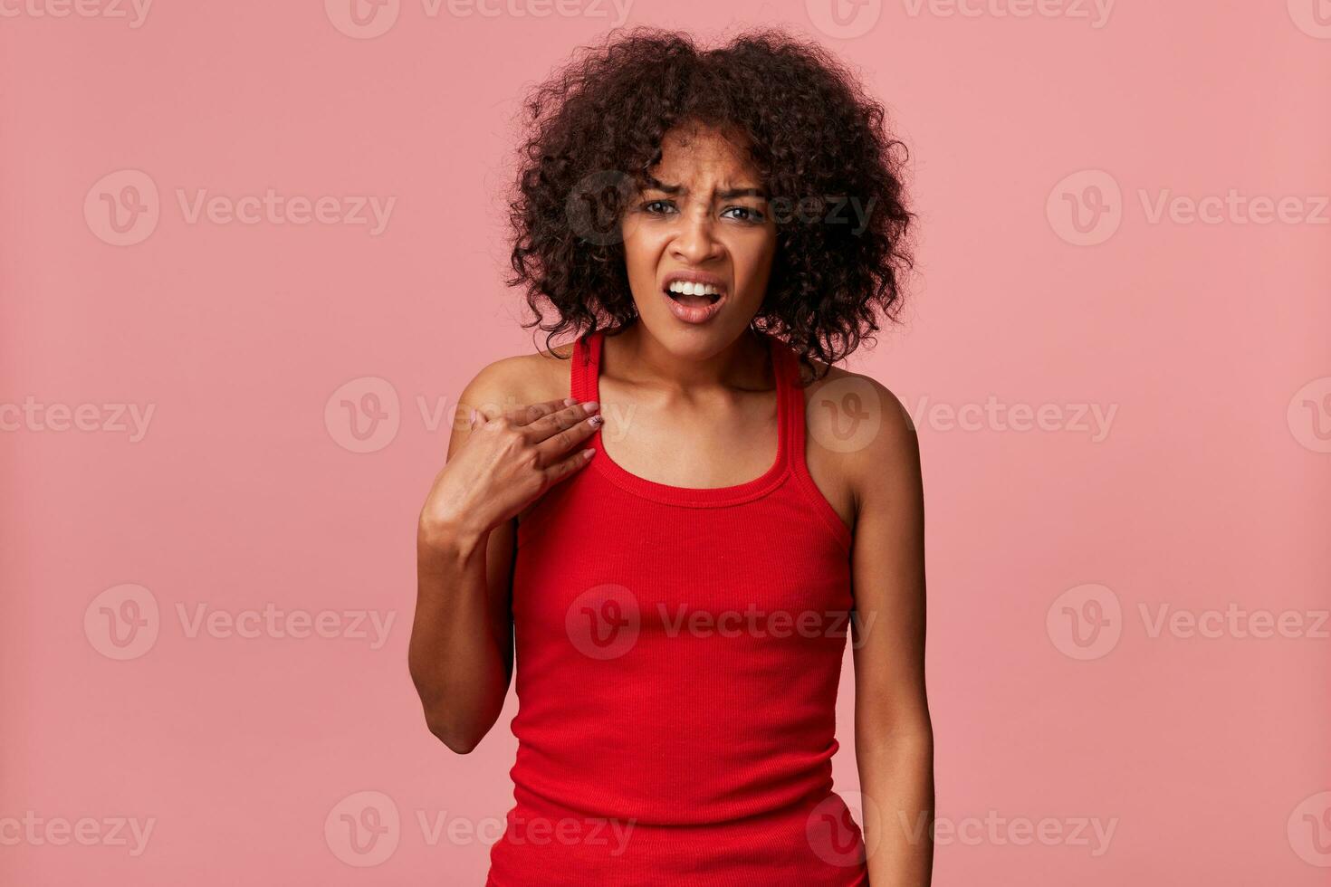 Angry outraged by an unjustified claim African American male wearing a red t-shirt, curly dark hair. Points with his left hand to herself, frowning and looking at camera isolated over pink background. photo