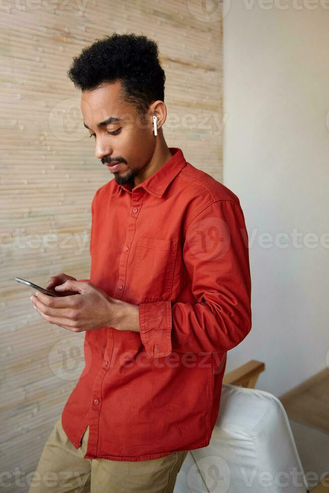 Concentrated young pretty bearded dark skinned brunette guy holding mobile phone and looking attentively on screen, posing over home interior in red shirt photo