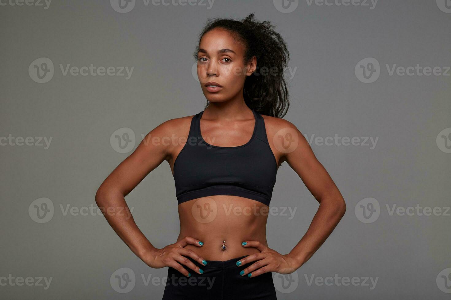 Studio shot of young pretty dark skinned curly lady with belly button piercing holding hands on her waist while posing over grey background in athletic black clothes photo