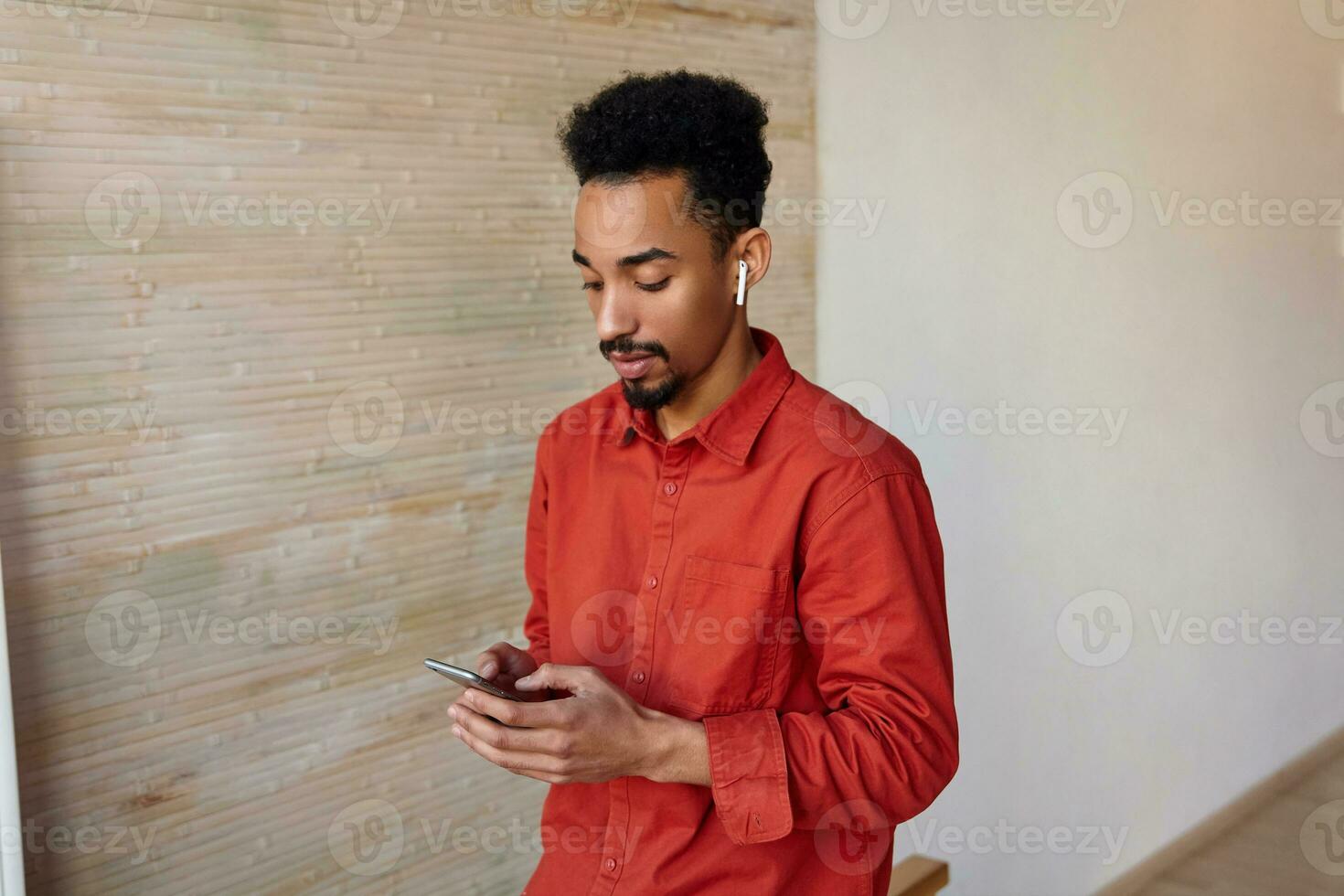 Indoor photo of young pretty bearded dark skinned man with short haircut wearing red shirt while standing over beige interior with smartphone in his hands