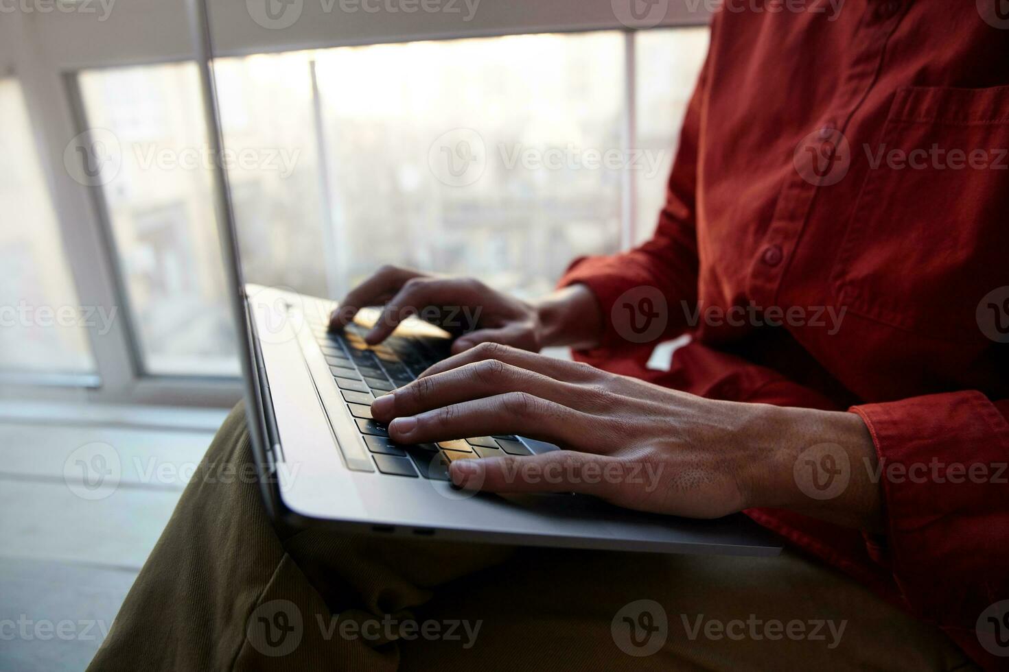 Cropped shot of young dark skinned guy dressed in red shirt and trousers keeping hands on keyboard while typing text, sitting near window on bright sunny day photo