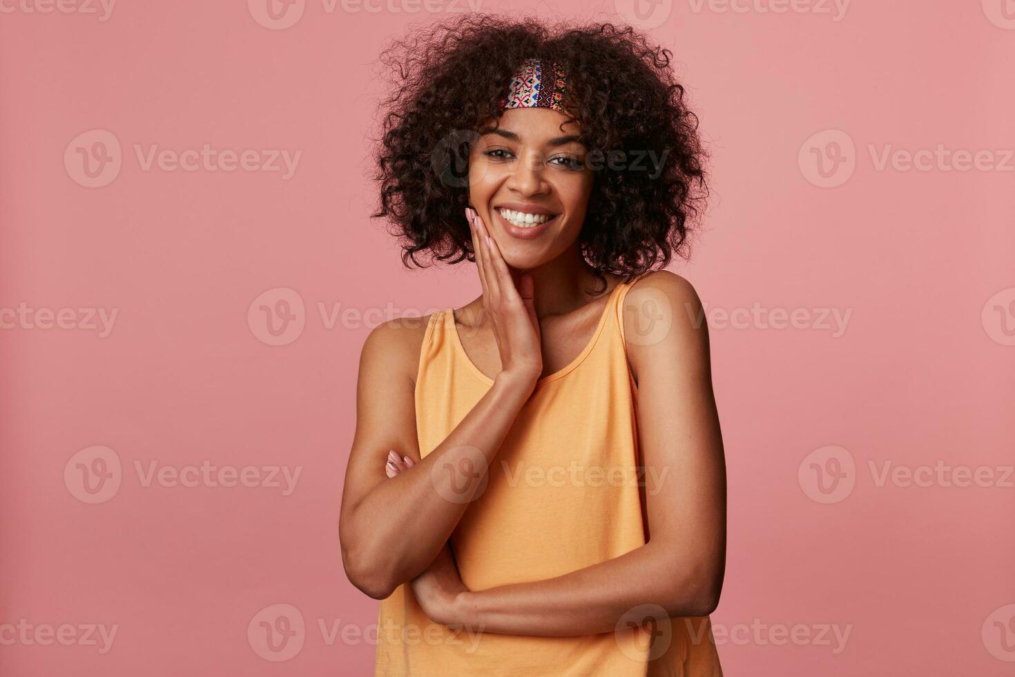 Happy pretty young curly brunette female with dark skin wearing casual hairstyle while standing over pink background, touching her face with raised palm and smiling sincerely photo