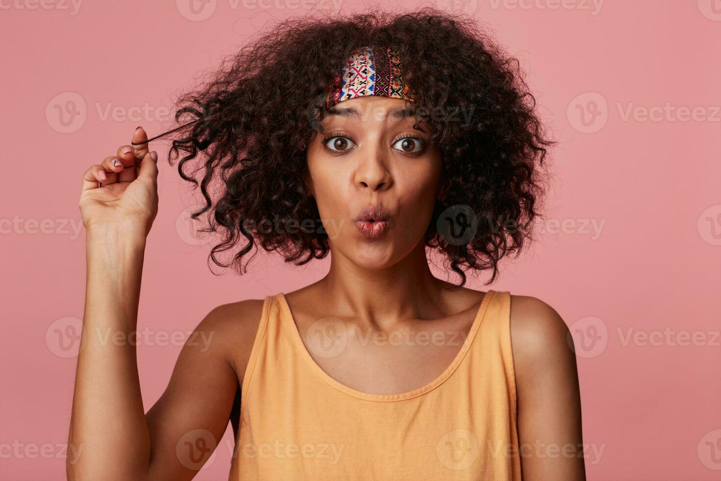 Portrait of charming young curly brunette female with dark skin looking surprisedly at camera and curling lips, playing with her short hair while standing over pink background in casual wear photo