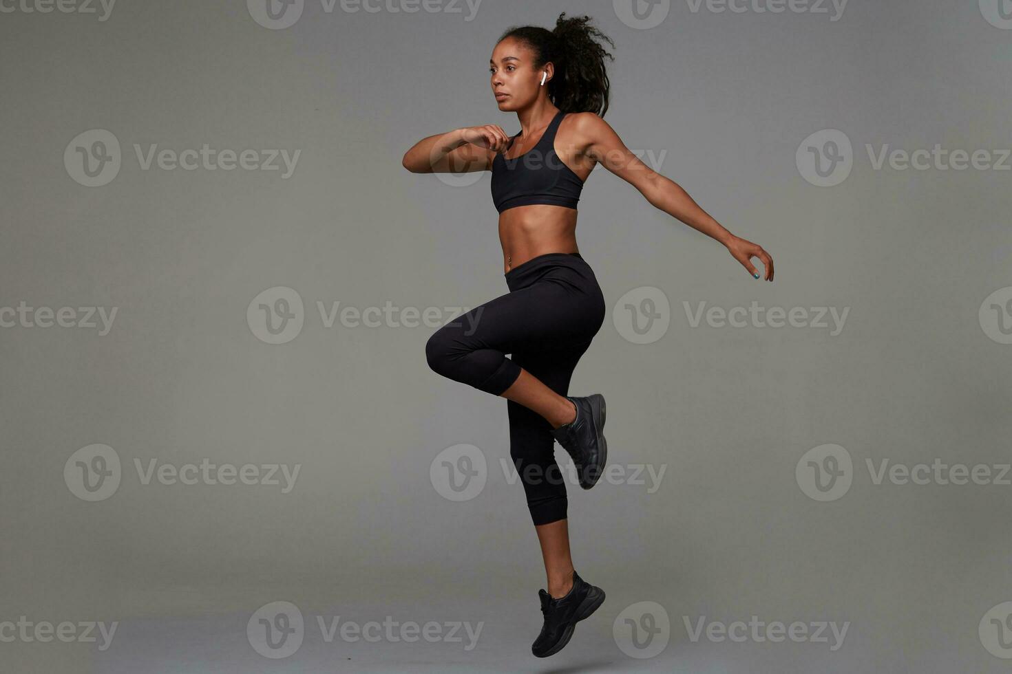 Full-length shot of slim young dark skinned curly brunette female being concentrated while making physical exercises in fitness studio, standing against grey background photo