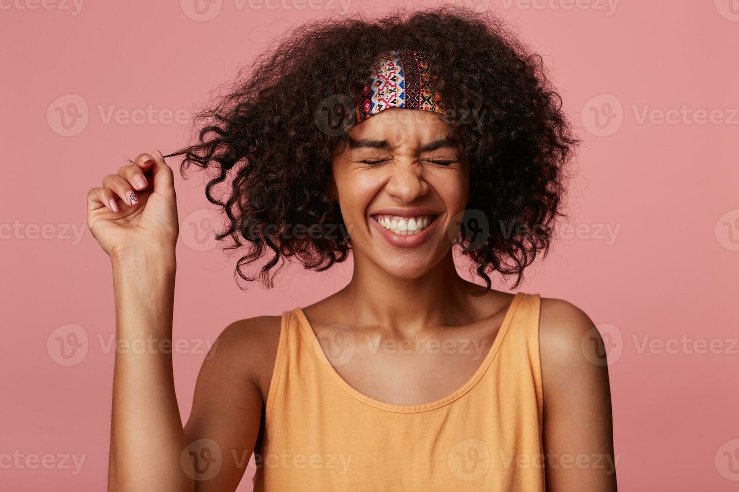 Pleasant looking happy dark skinned curly brunette woman with casual hairstyle smiling widely with closed eyes while standing over pink background in colorful headband and light orange shirt photo