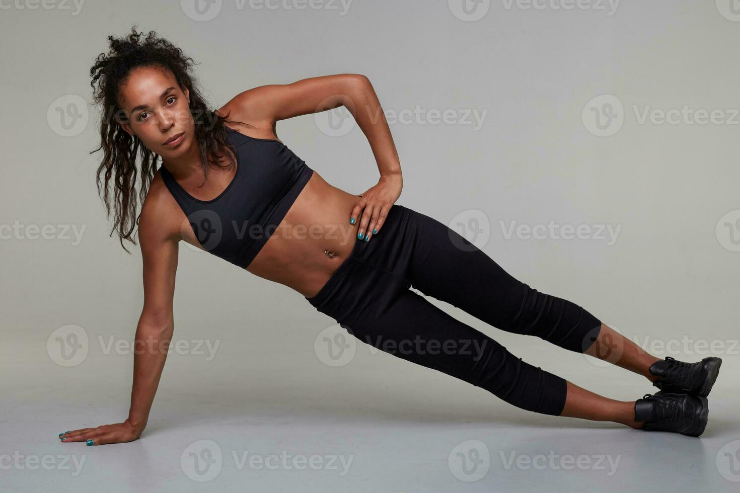 Horizontal photo of young sporty dark skinned woman with curly long hair making physical exercises in gym class, wanting to be fit and healthy, isolated over grey background