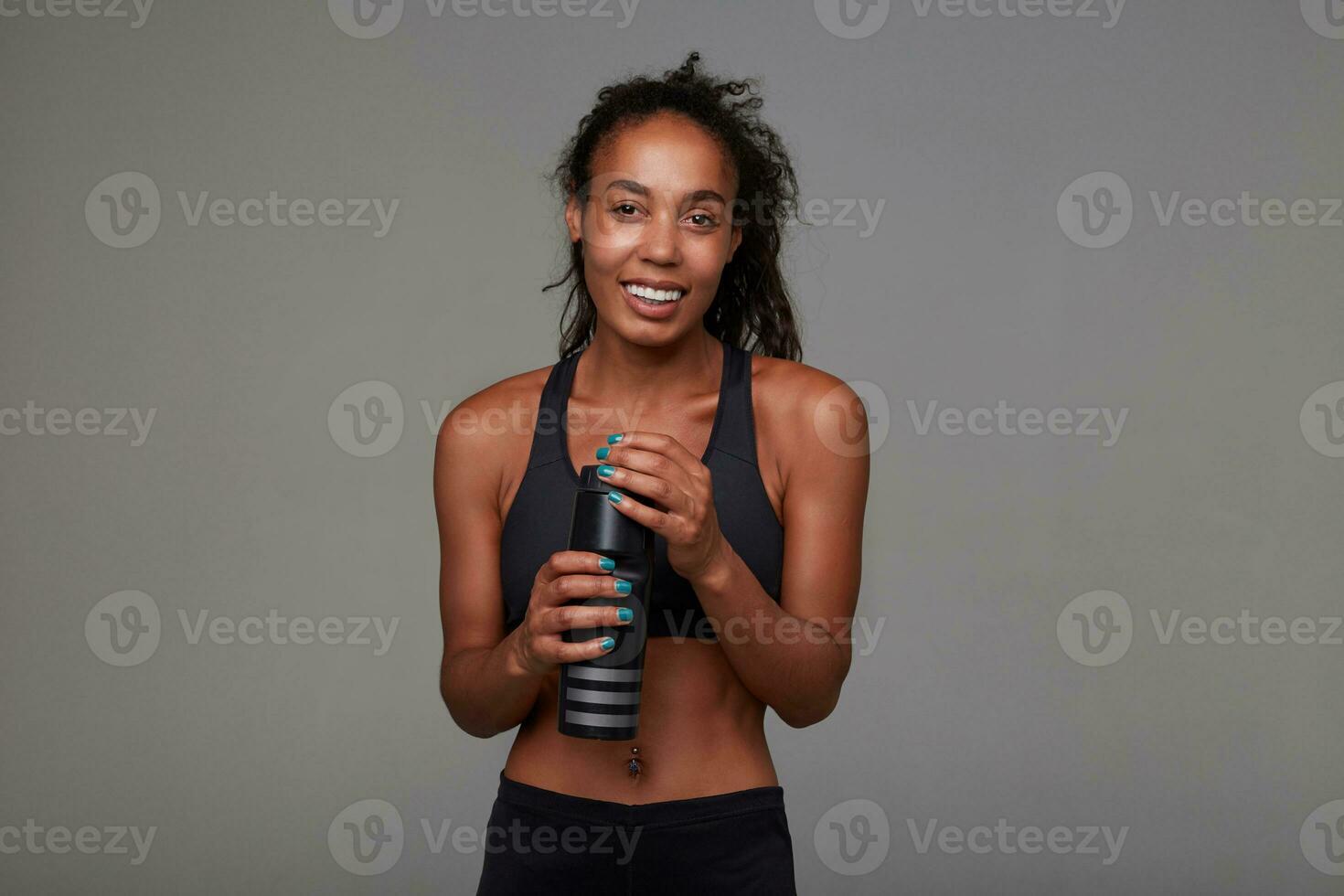 Indoor photo of happy young athletic dark skinned brunette female having break from fitness and drinking water while standing over grey background, being in nice mood