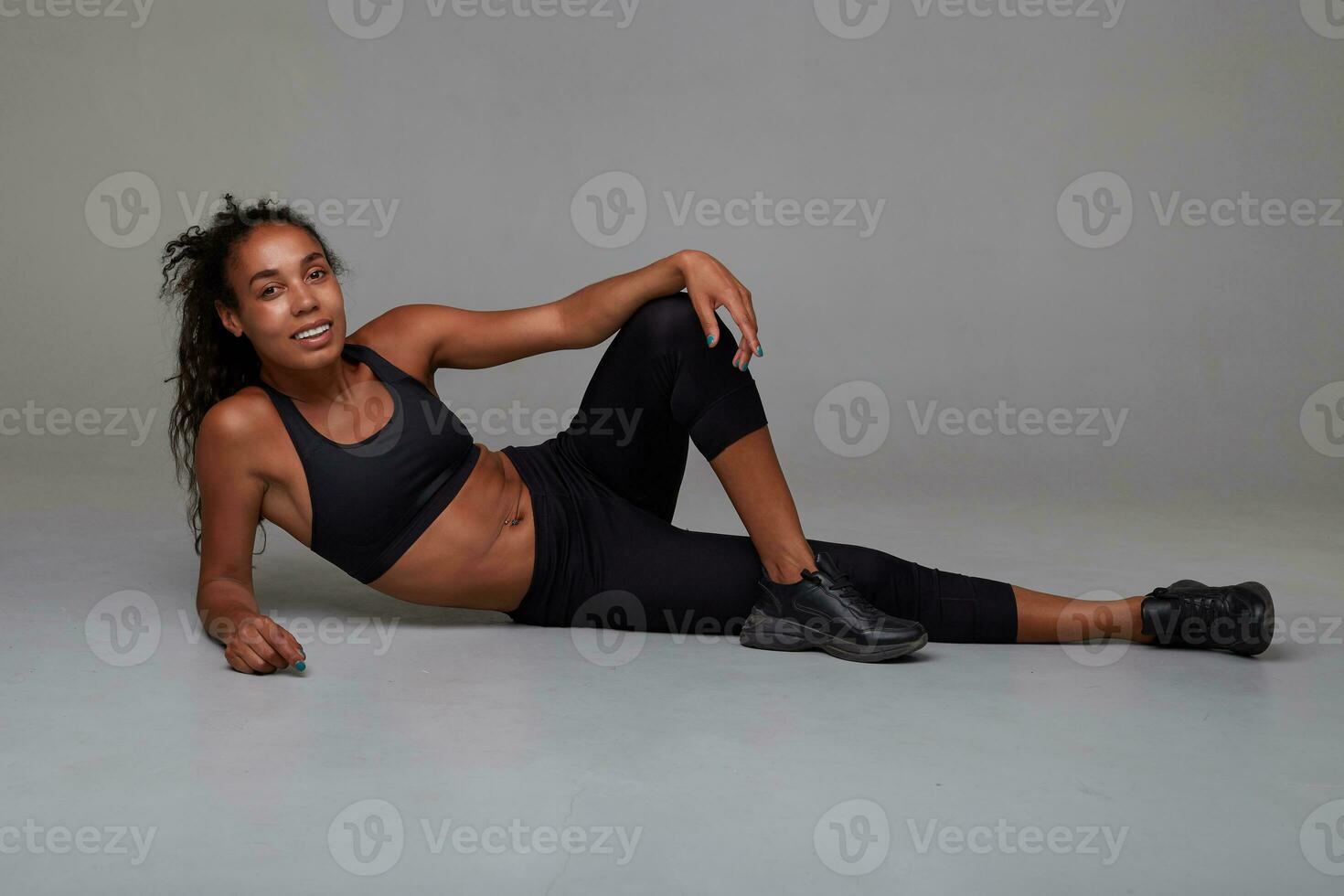 Full-length photo of sporty young dark skinned curly lady making small beak between workout sessions, lying on floor over grey background and smiling widely to camera