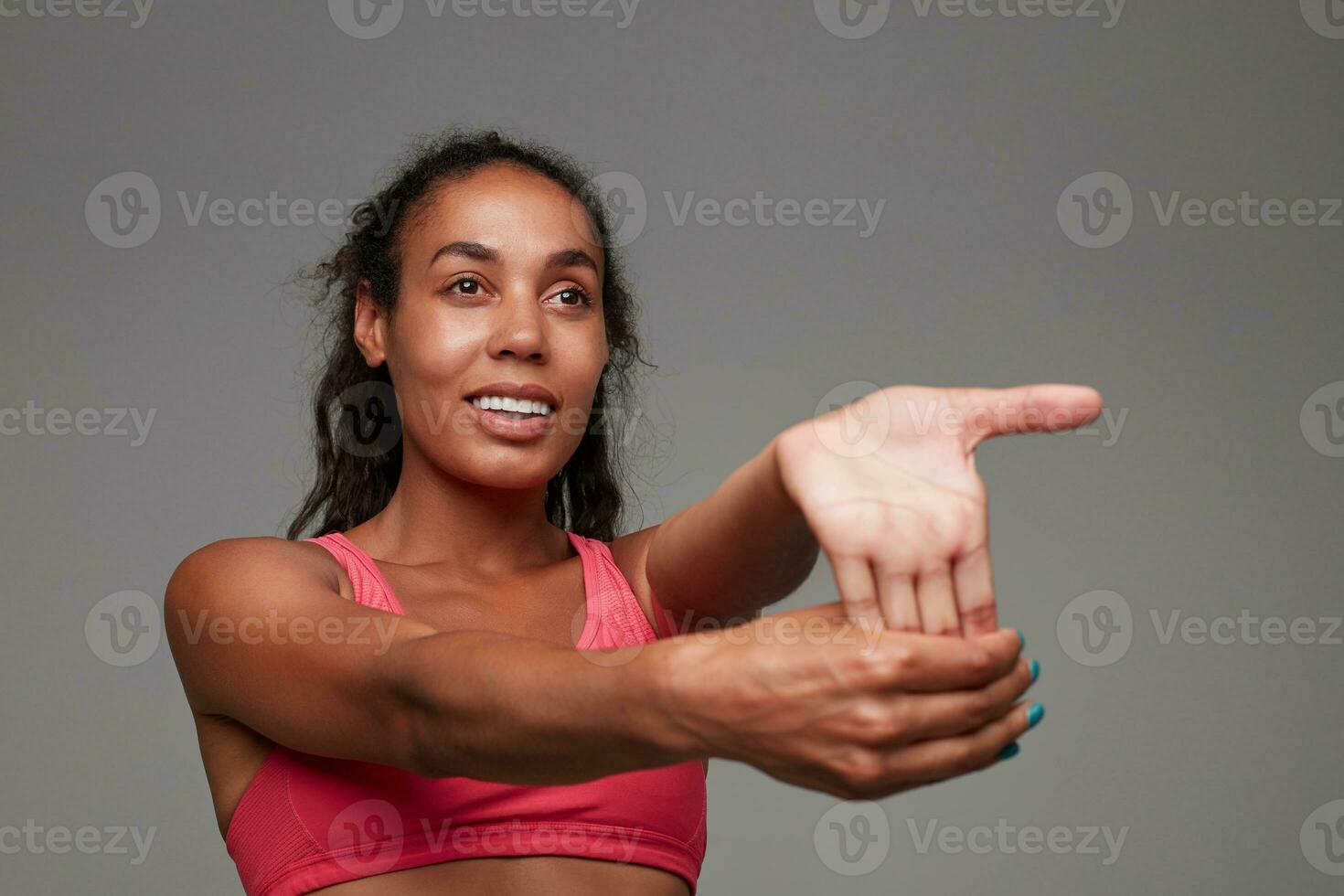 Portrait of attractive young dark skinned curly brunette woman dressed in athletic pink top while standing over grey background, looking positively ahead while stretching her arm photo
