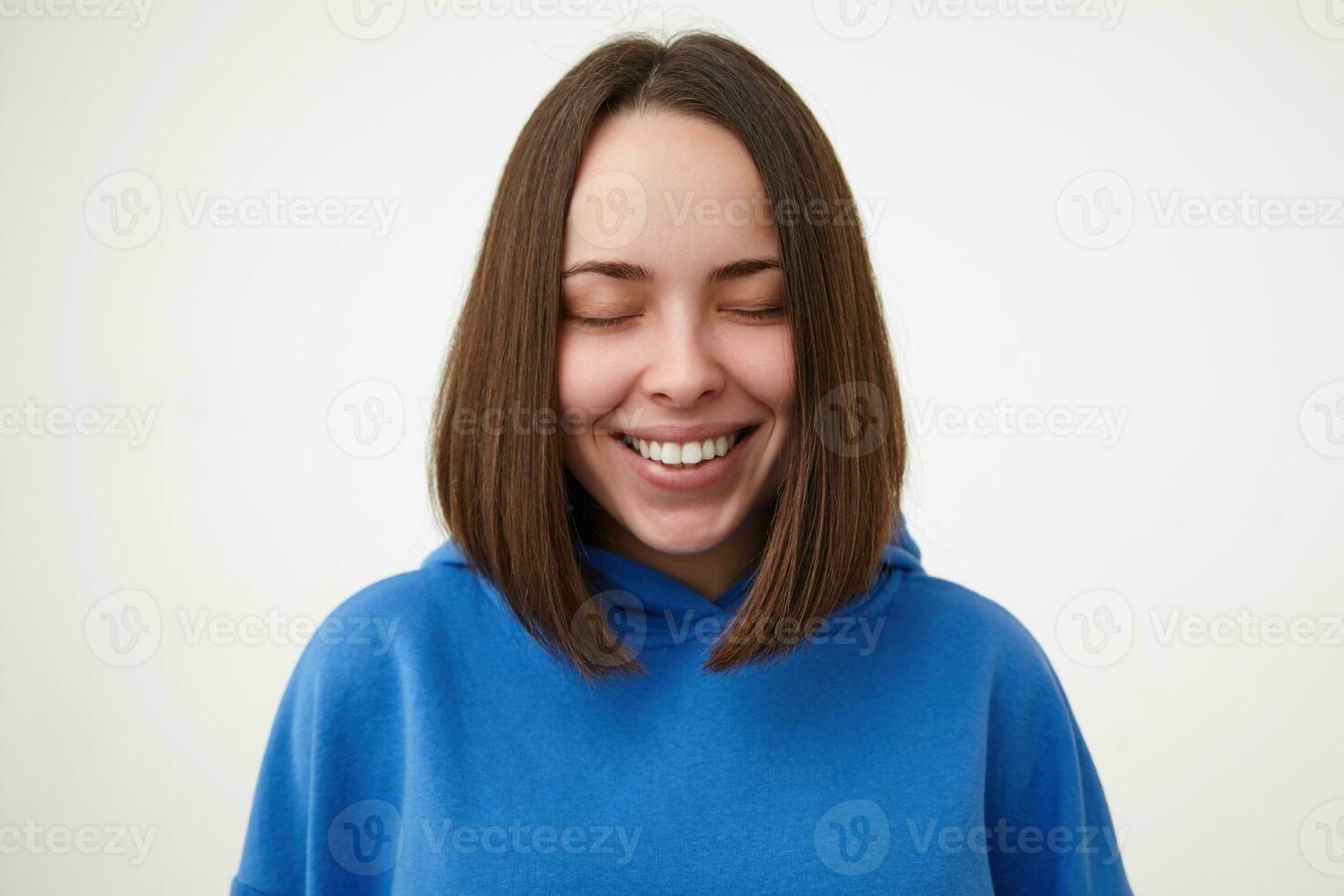 Pleasant looking young cheerful brown haired woman with bob haircut keeping her eyes closed while smiling happily, dressed in sporty clothes while posing over white background photo
