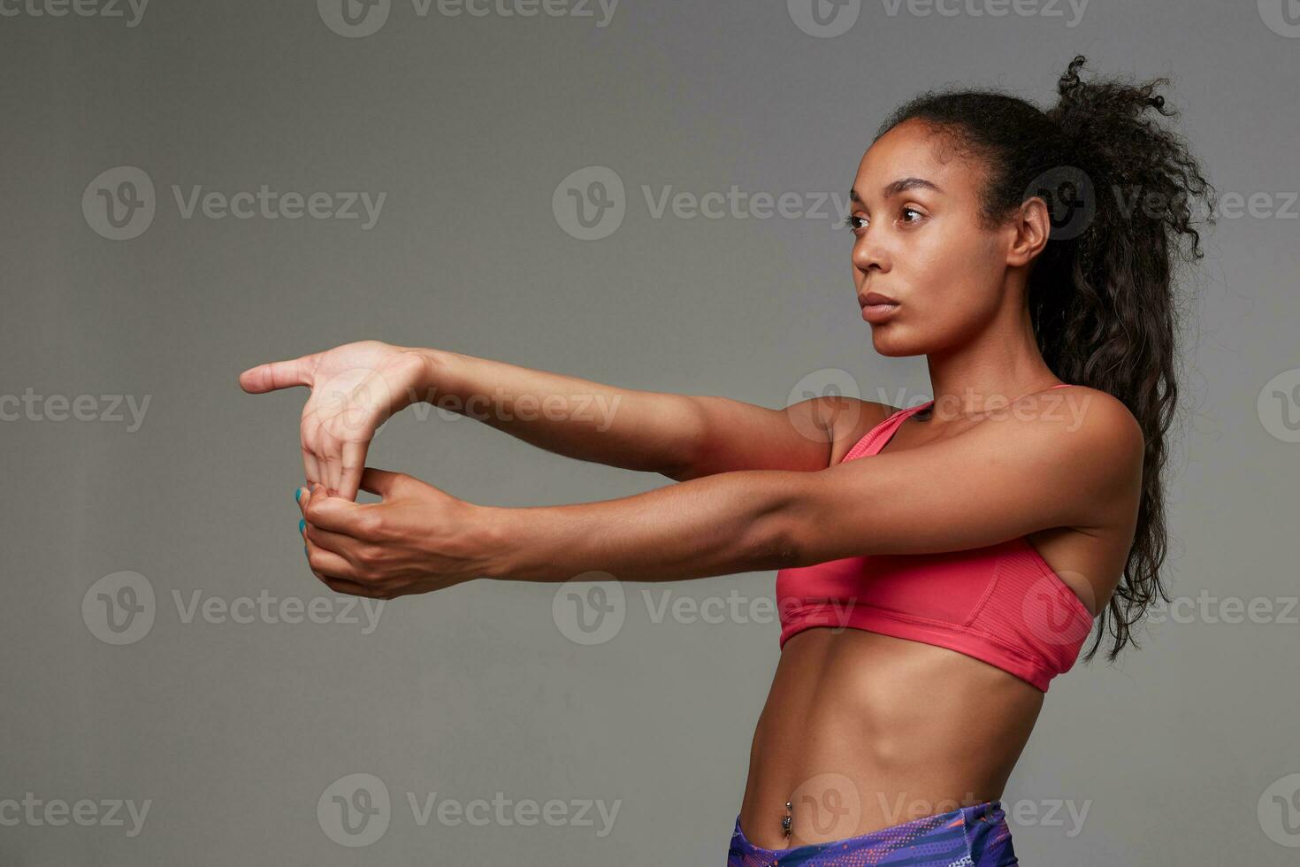 espalda ver de joven deportivo oscuro desollado Rizado largo peludo morena mujer extensión su brazos después gimnasia, mirando en frente de su con concentrado rostro, aislado terminado gris antecedentes foto