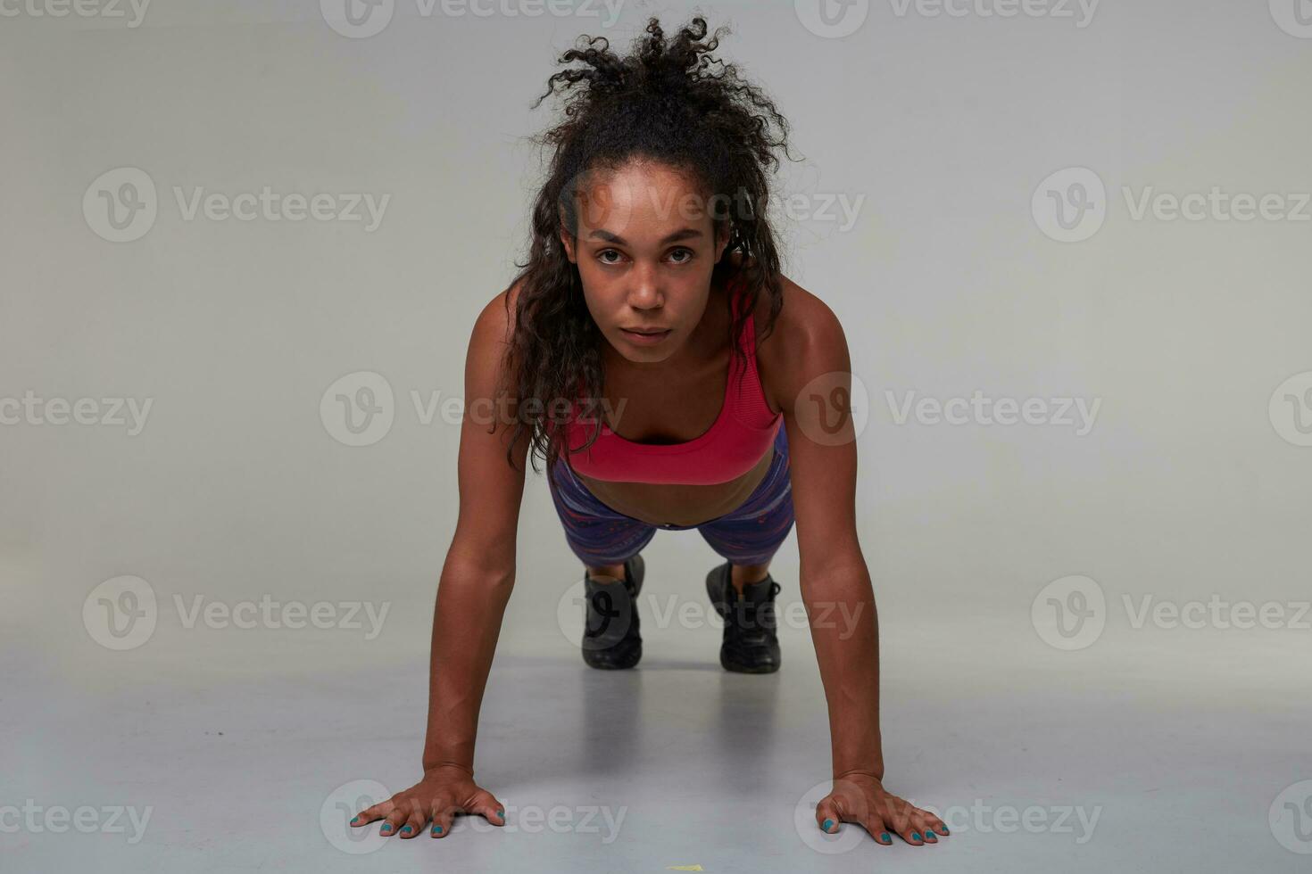 Sporty young dark skinned curly brunette female with casual hairstyle making plank exercise indoor and looking at camera with concentrated face, isolated over grey background photo