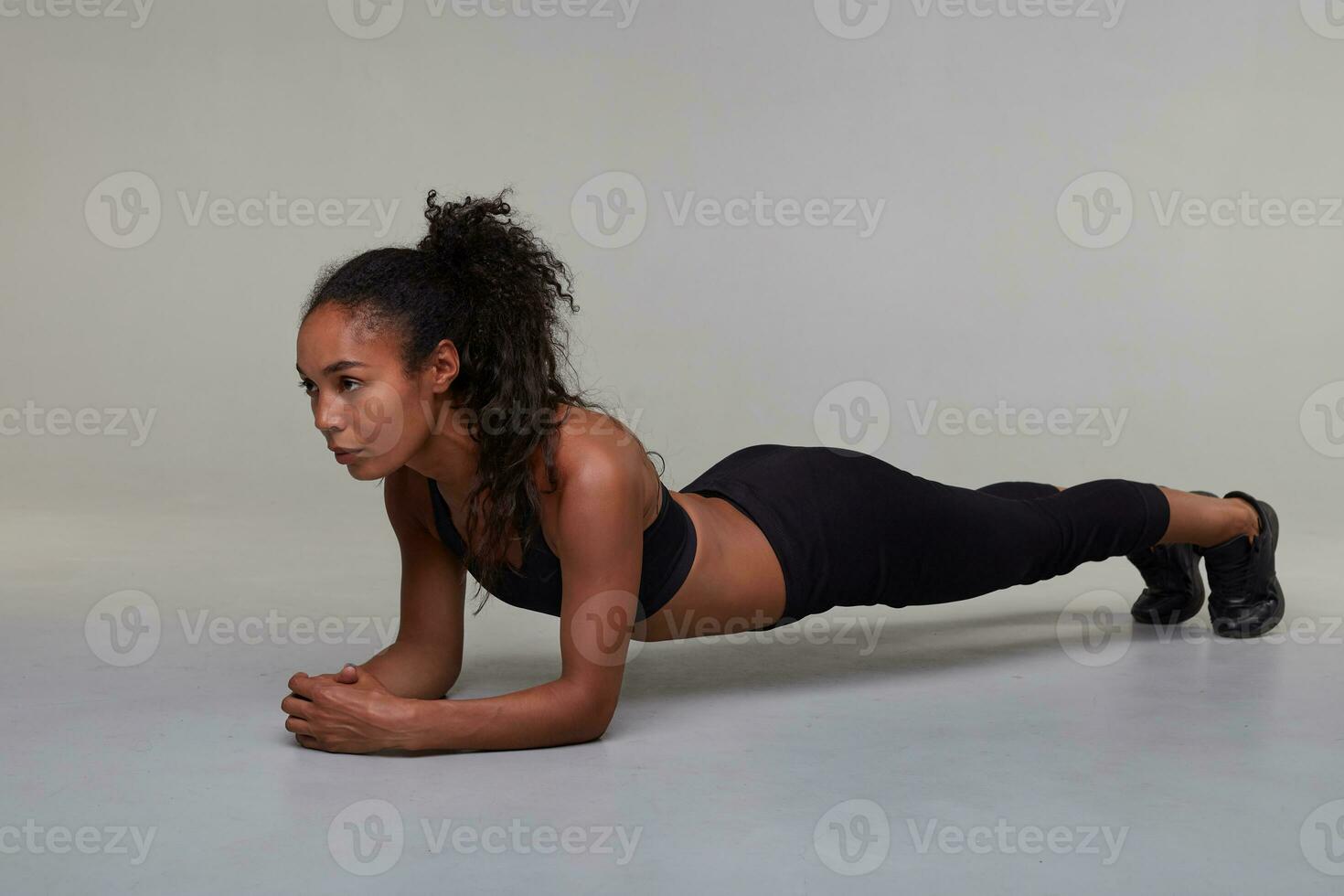 Indoor shot of young slim dark skinned curly brunette woman going for sports before work, planking and looking in front of herself intently, standing against grey background photo