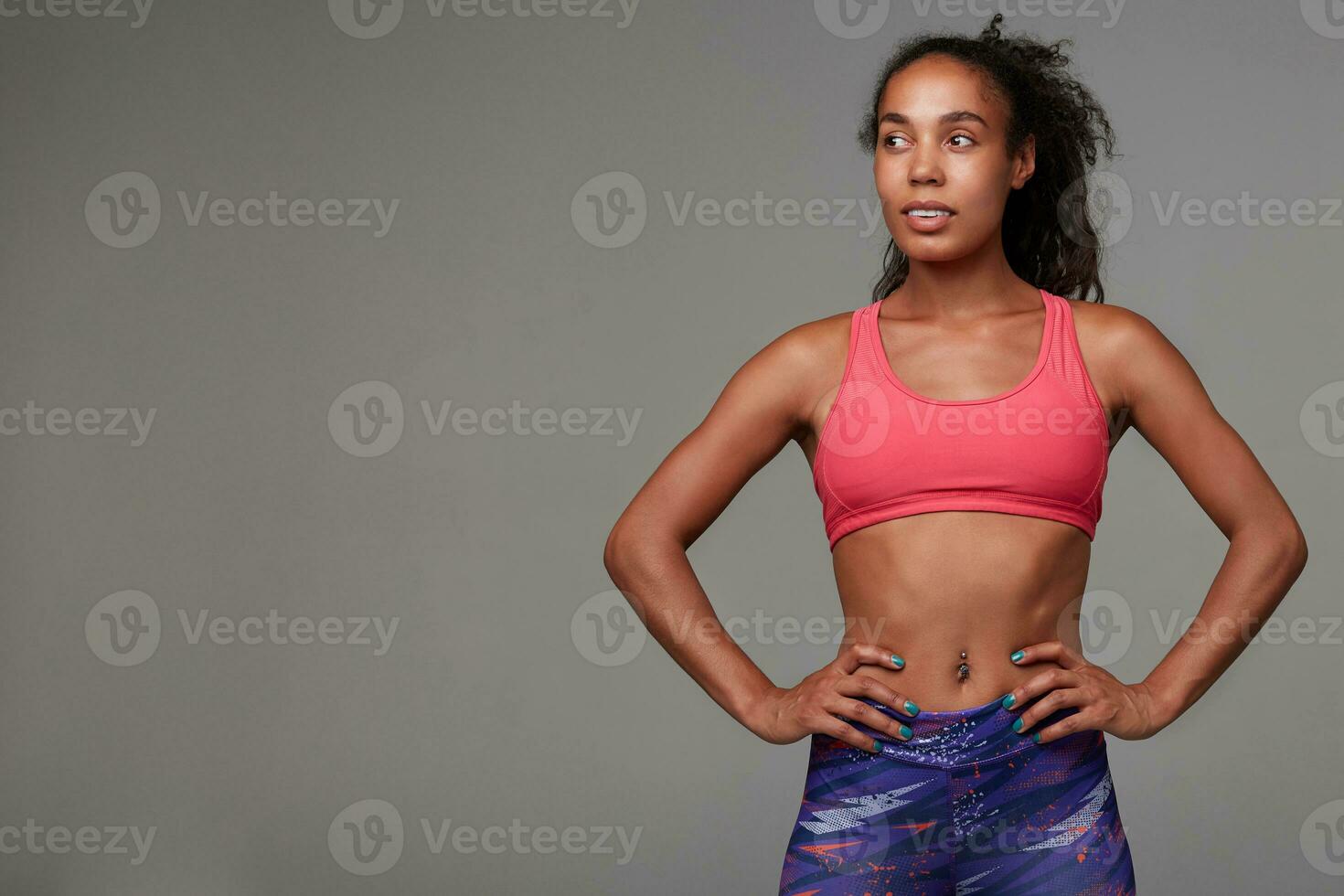 Indoor photo of pretty young dark skinned brunette woman dressed in athletic clothes looking positively aside and smiling slightly, standing against grey background