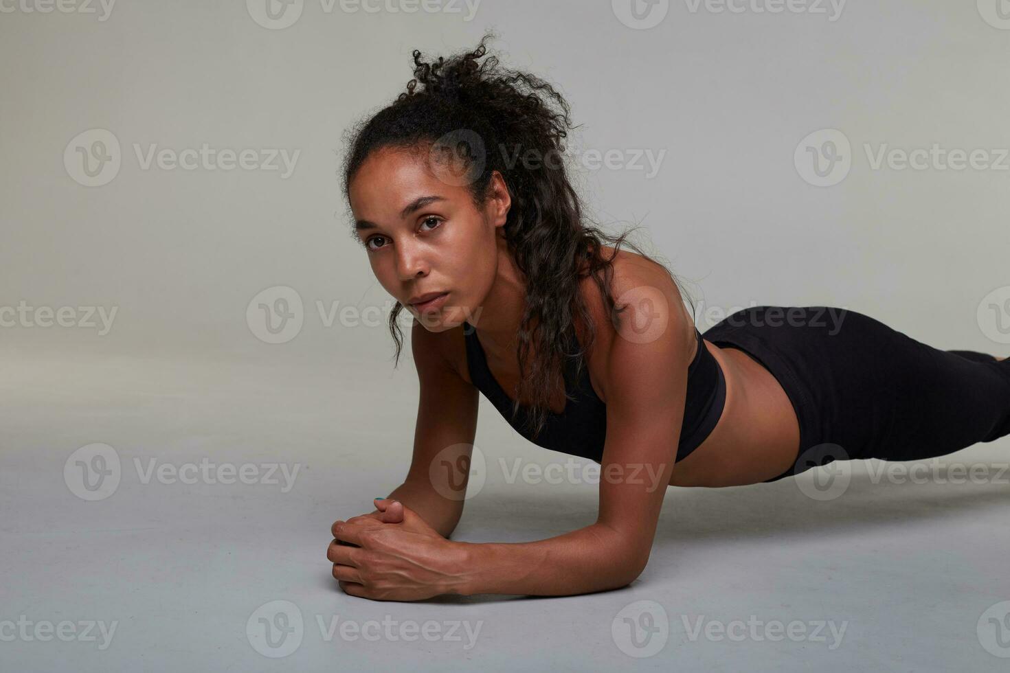 Portrait of young dark haied concentrated curly lady staying in plank while posing over grey background, looking thoughtfully aside with folded lips and keeping arms crossed photo