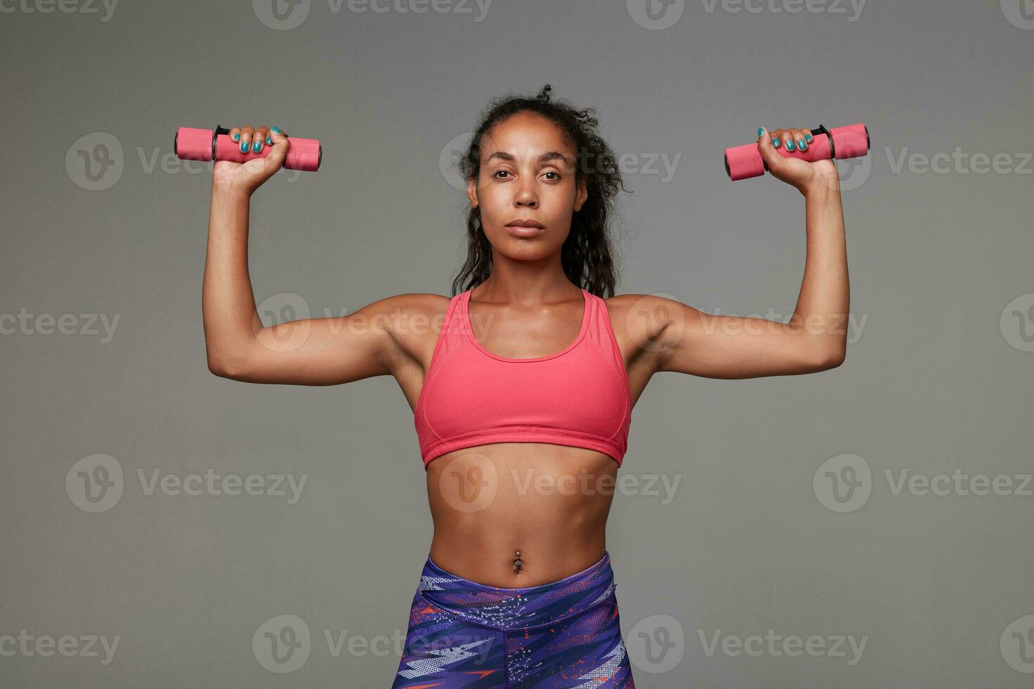 Indoor photo of athletic dark skinned woman with curly long brown hair doing workout for her hands while standing over grey background, dressed in sporty clothes