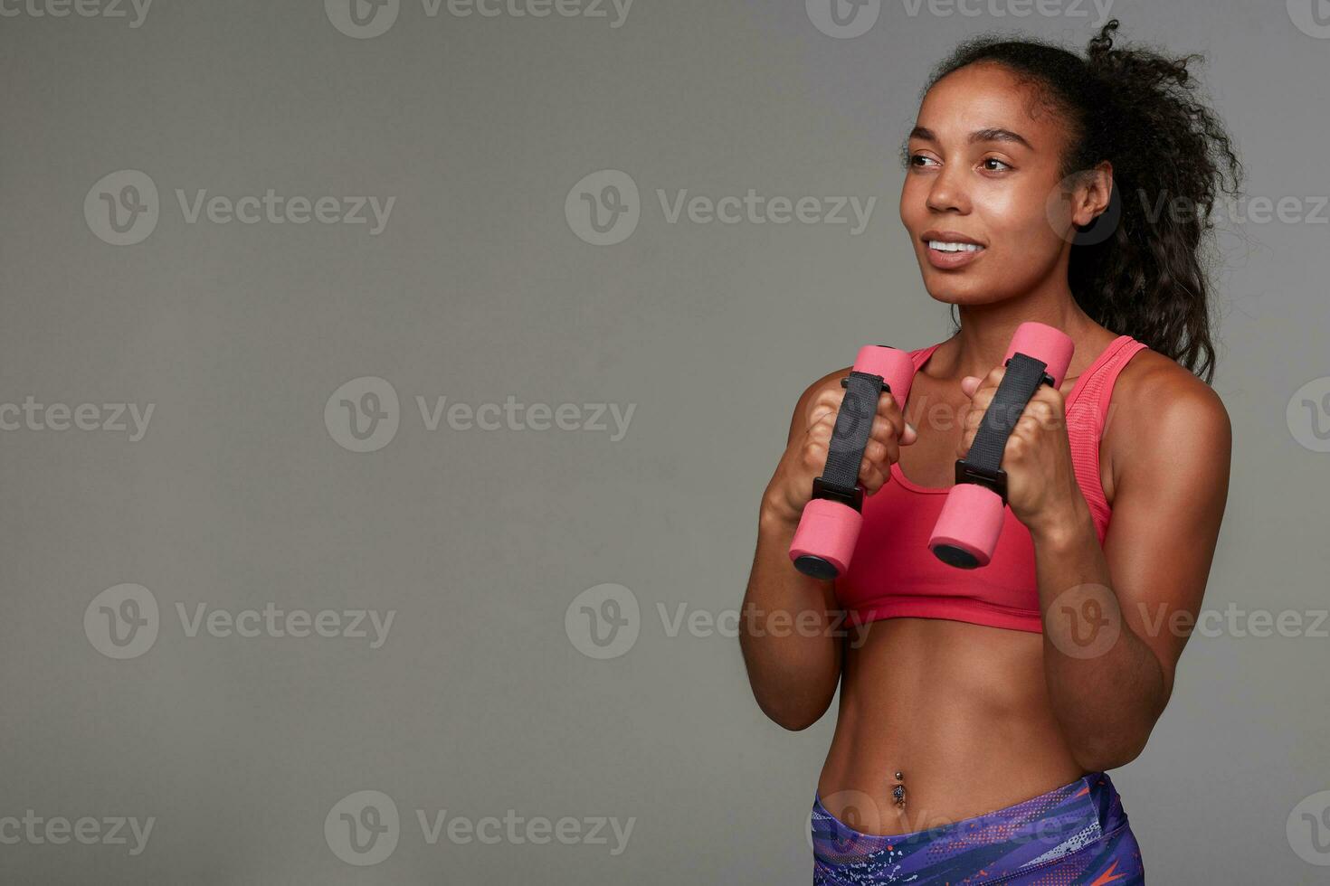 Cheerful pretty young curly long haired brunette lady with dark skin doing morning exercises with dumbbells and looking positively ahead, isolated over grey background photo