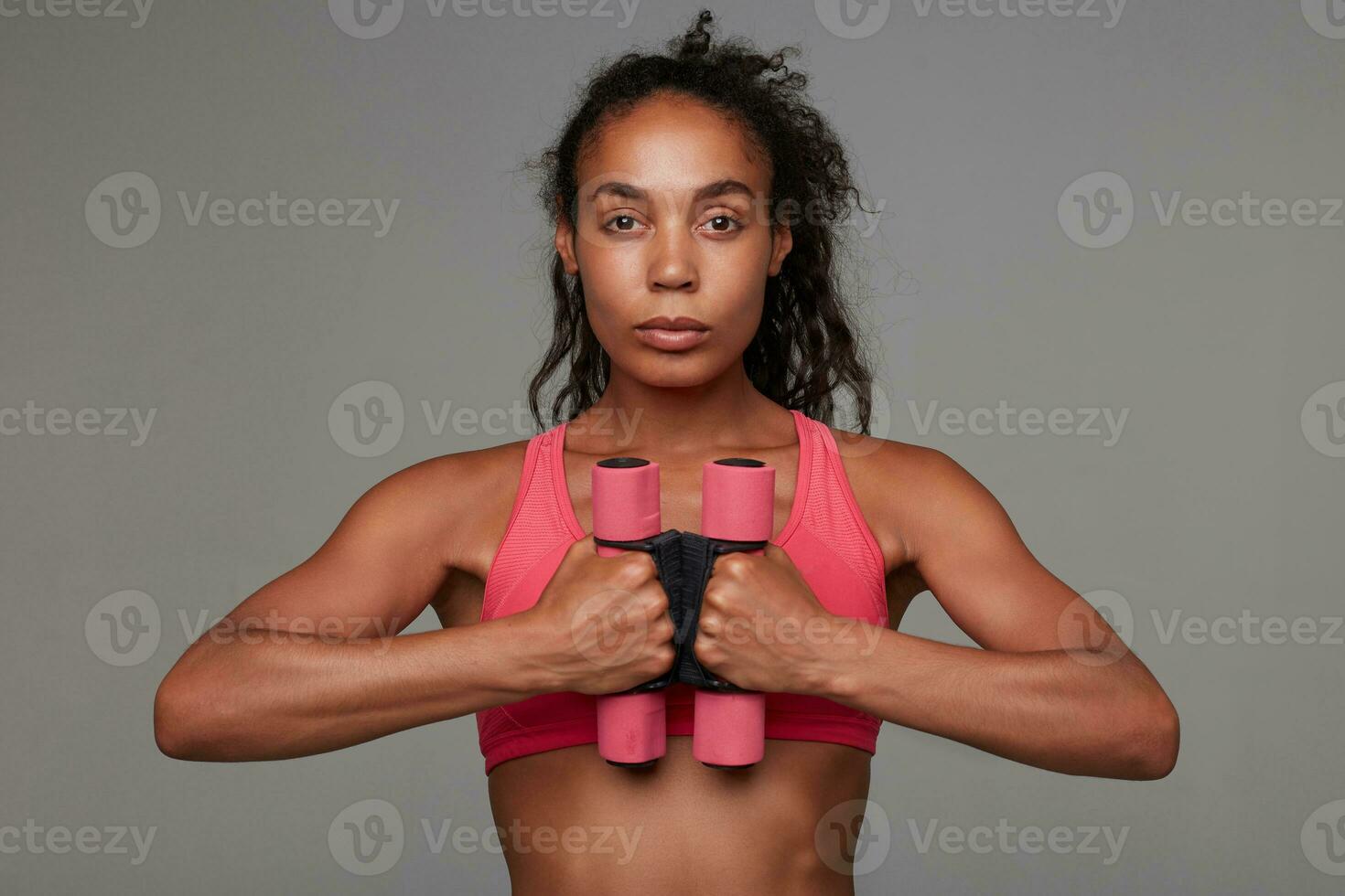 Portrait of young athletic brown-eyed dark skinned woman with casual hairstyle holding weighting agents at chest level and looking calmly at camera with folded lips, isolated over grey background photo