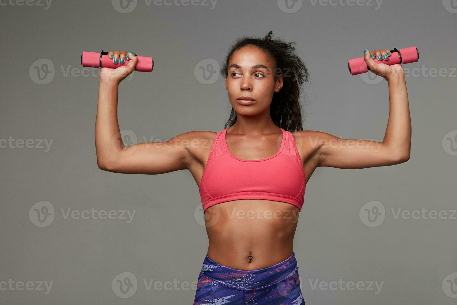 Fitness young slim dark skinned curly brunette female looking aside with folded lips while making exersices with dumbbells, goes in for sport every morning, isolated over grey background photo