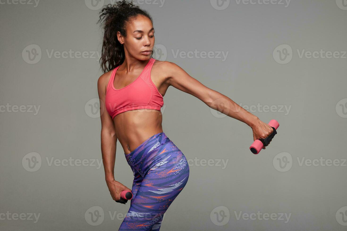 Fitness young slim dark skinned curly woman in pink sporty top and printed leggins doing workout for her hands while standing against grey background. Sport and wellness concept photo