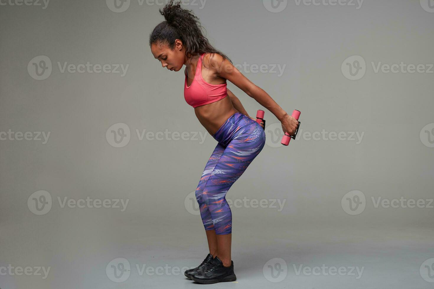 Side view of sporty young dark skinned curly long haired woman dressed in athletic clothes holding dumbbells behind her back while making exercises for hands, isolated over grey background photo