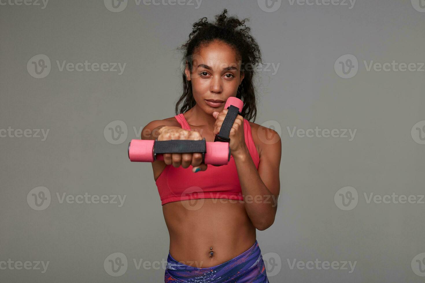 Fitness young dark skinned model with curly long brown hair making exercises with dumbbells while posing over grey background. Sport and healthy lifestyle concept photo