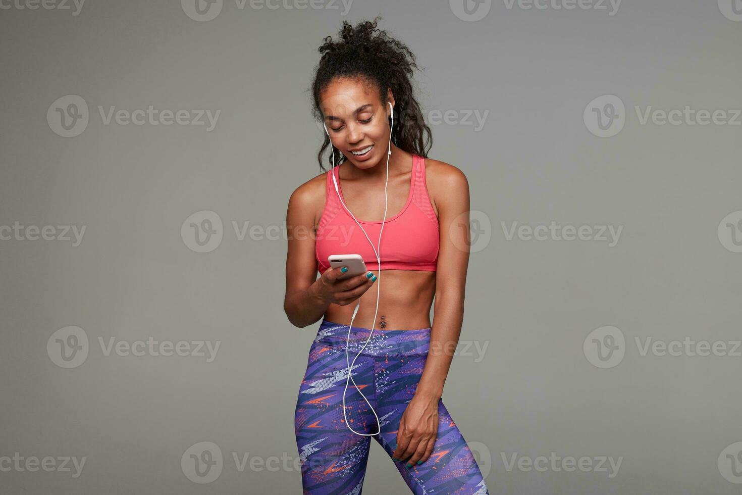 Good looking young lovely dark skinned curly brunette sportswoman with belly button piercing holding smartphone in raised hand and smiling happily, isolated over grey background photo