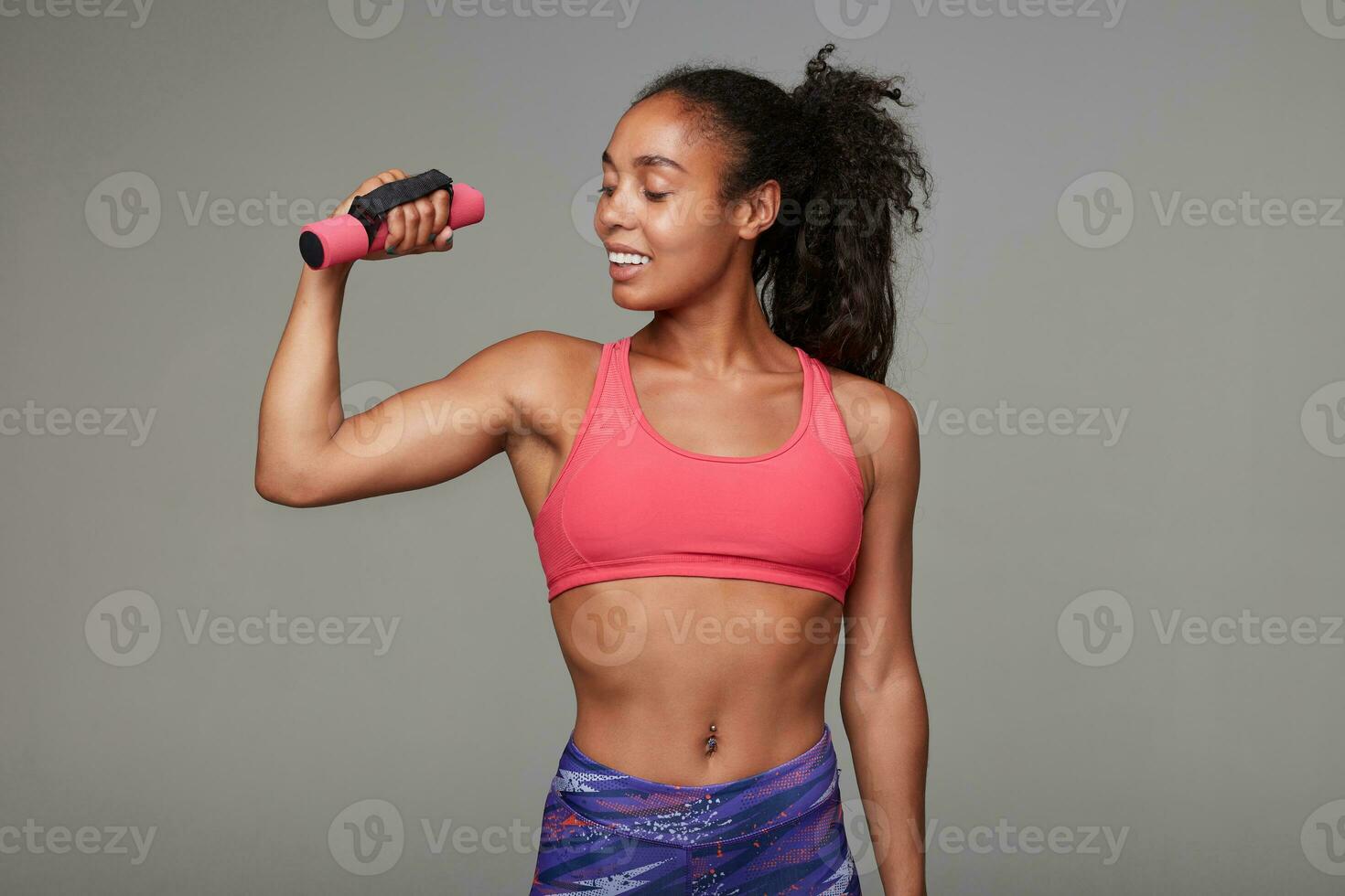 Positive sporty young dark skinned curly lady with ponytail hairstyle looking happily on her raised hand, being proud of her strong biceps, isolated over grey background photo