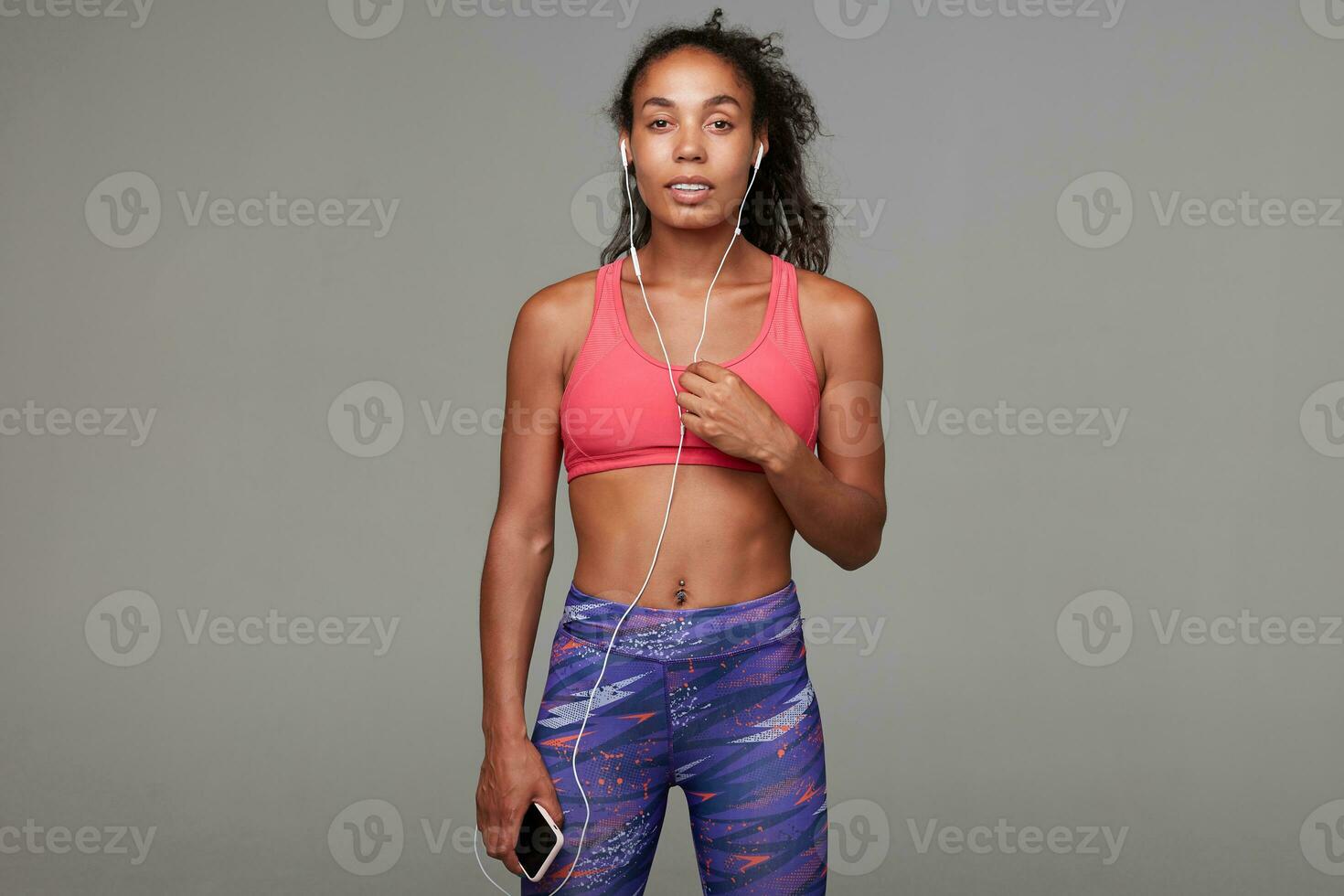 Studio photo of young pretty dark skinned female with curly brown hair being in good physical form, making break with training and enjoying music in her headphones over grey background