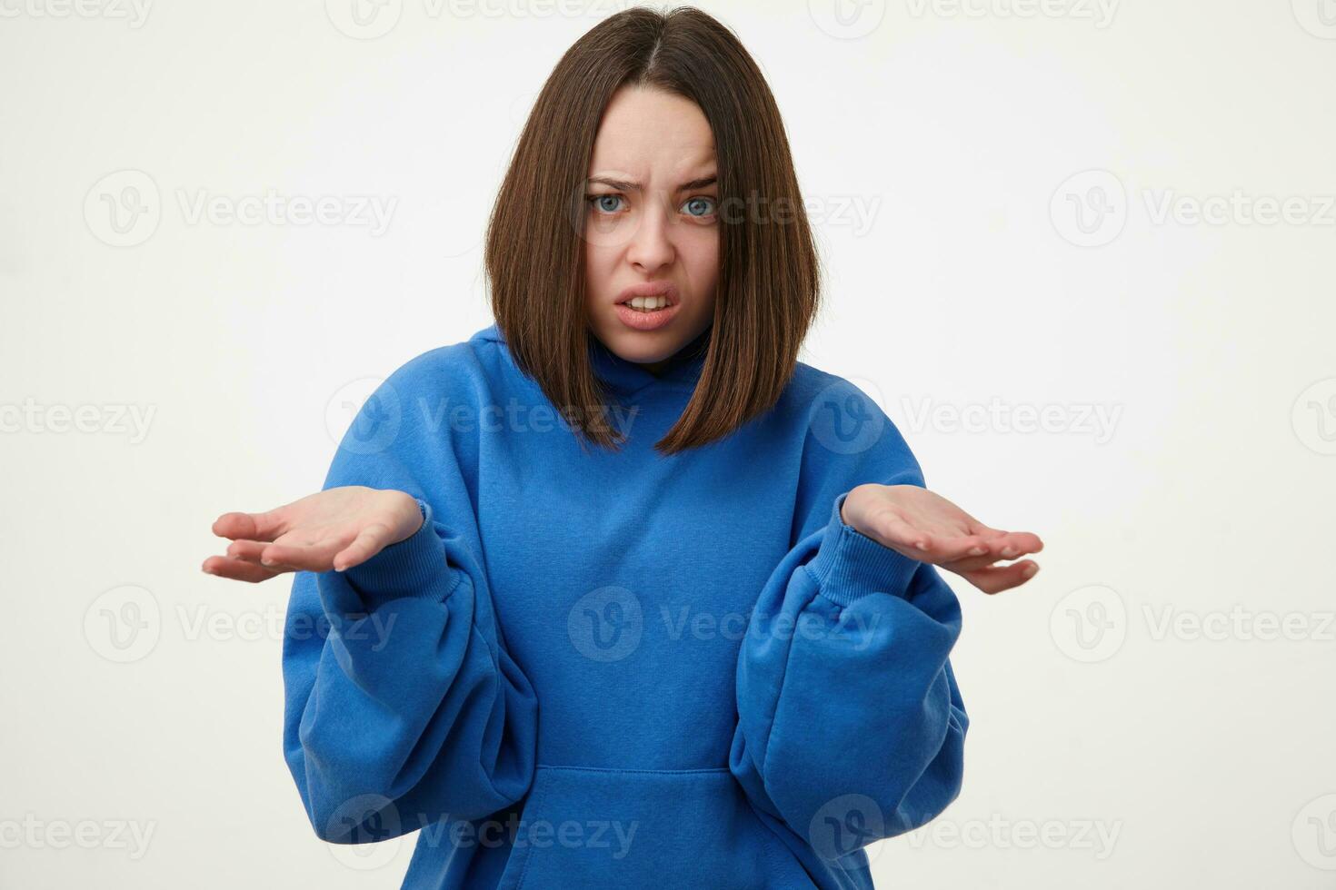 Bewildered young short haired brunette female without makeup grimacing her face and keeping palms raised while standing over white background in sporty wear photo