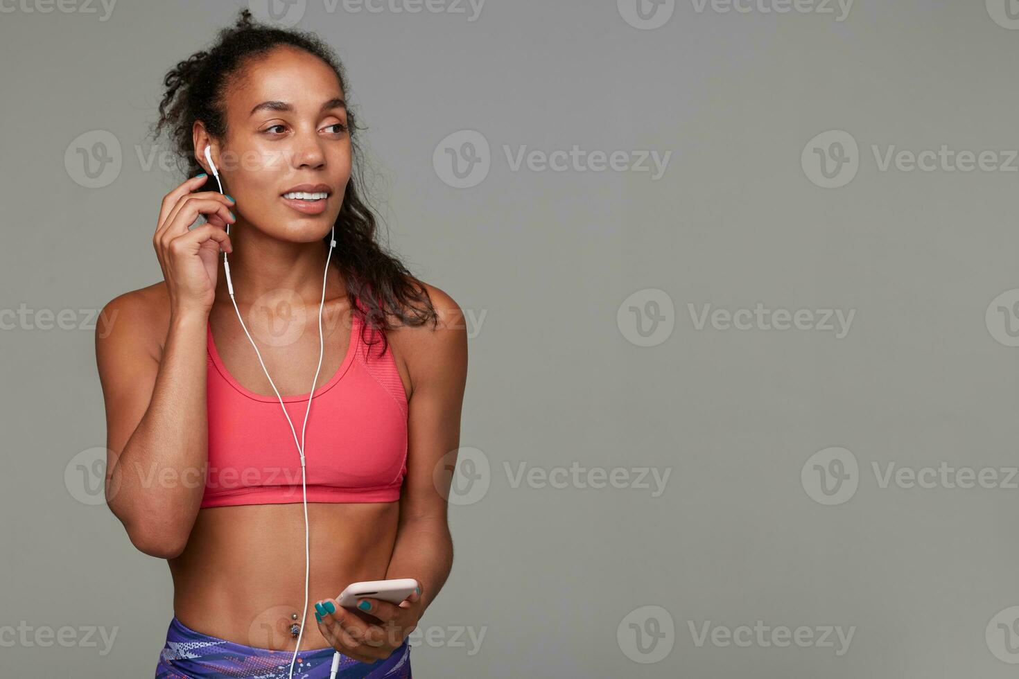 Studio shot of young slim curly long haired brunette lady with dark skin inserting earpiece into her ear and looking aside with pleasant smile, relaxing after gym over grey background photo