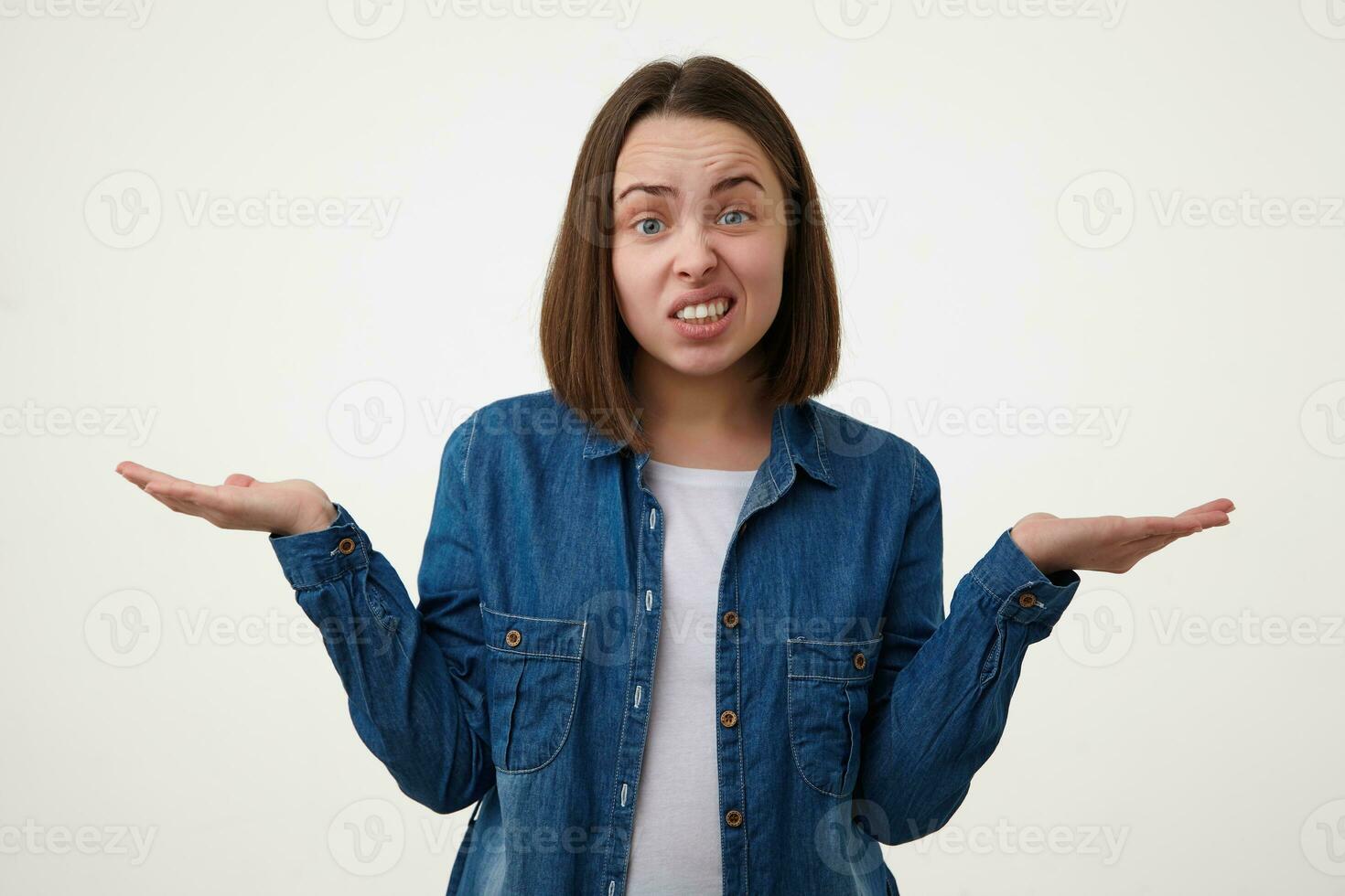 Indoor shot of bewildered young brown haired female with short haircut grimacing confusedly her face while standing over white background with raised palms photo