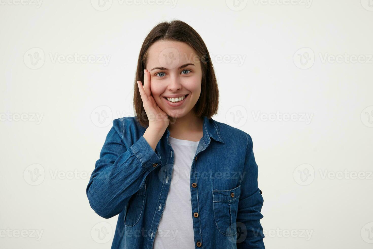 Indoor photo of pleased young short haired brunette female touching gently her face with raised hand and looking gladly at camera with charming smile, isolated over white background