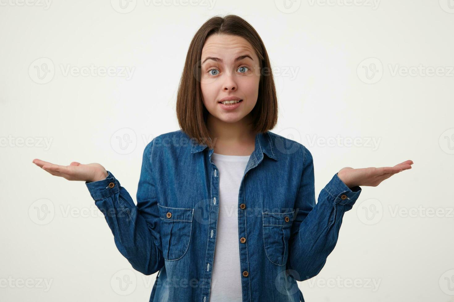 Puzzled young lovely blue-eyed brunette woman with bob haircut raising confusedly hands while looking surprisedly at camera, standing over white background photo