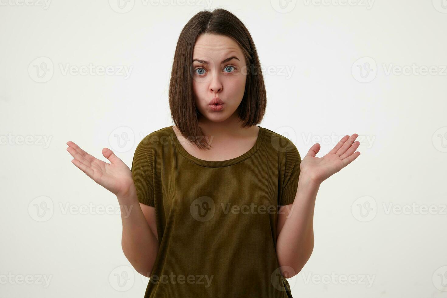 Bewildered young pretty short haired brunette female raising confusedly hands and pouting her lips while looking at camera, isolated over white background photo