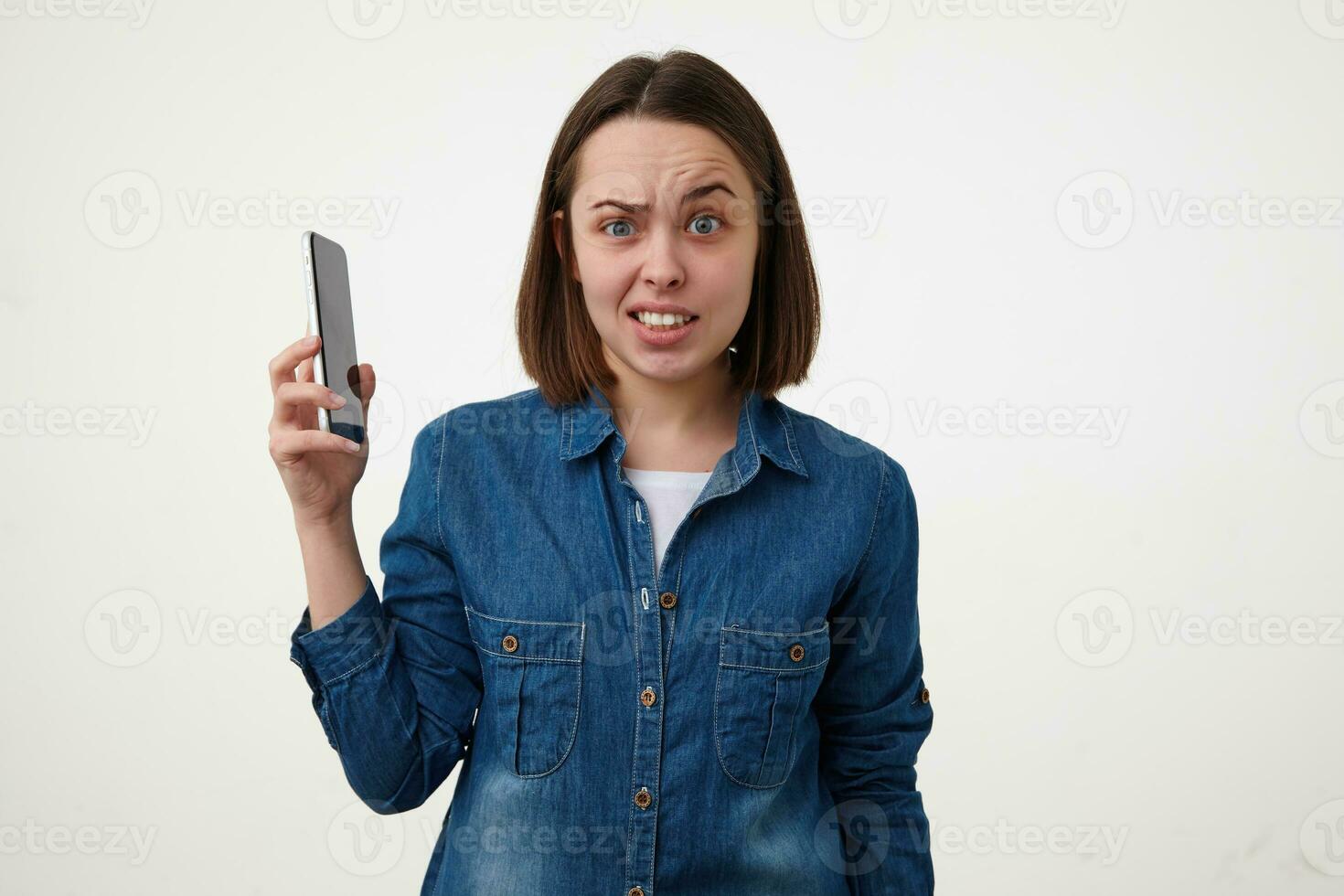 Studio shot of bewildered young brunette female raising confusedly her eyebrow while looking at camera and raising hand with smartphone in it, isolated over white background photo