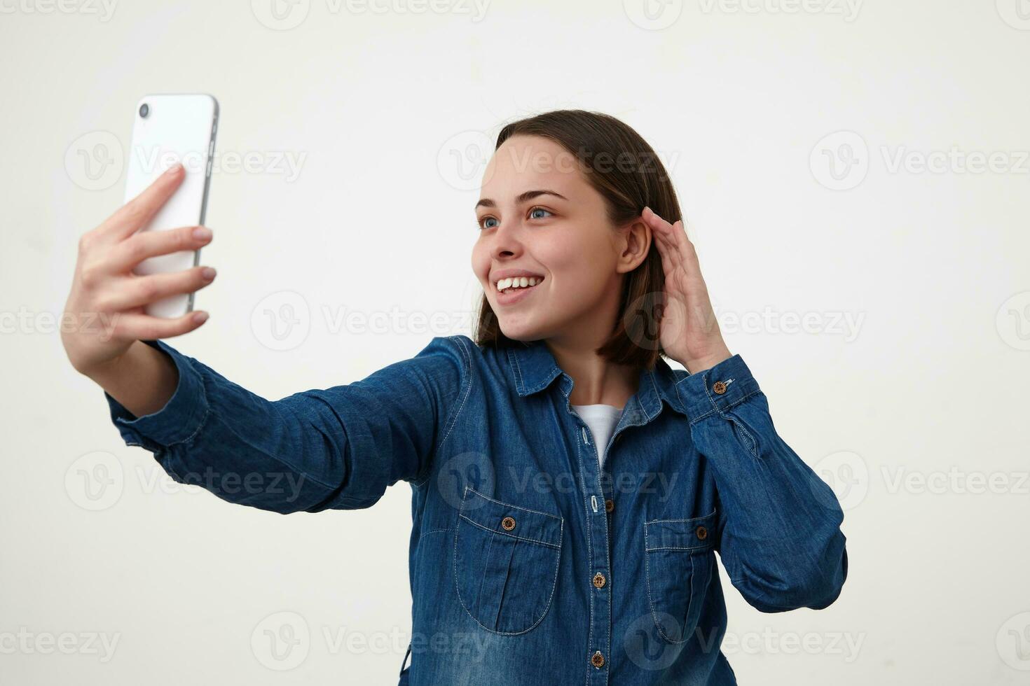 Positive young lovely short haired brunette lady smiling nicely while making photo of herself, dressed in white t-shirt and blue jeans coat while posing over white background