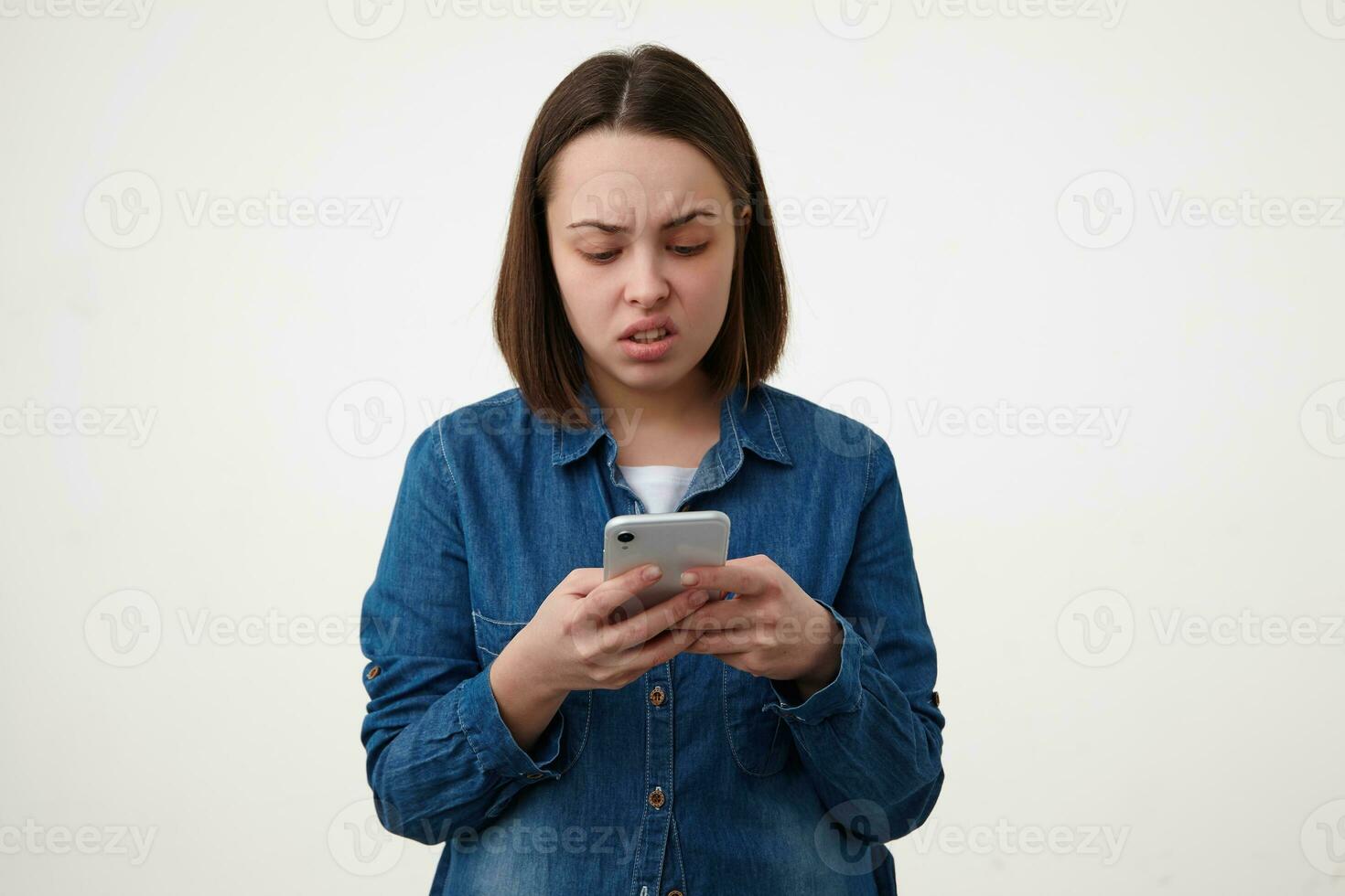 Studio shot of confused young brunette lady with short haircut twisting her mouth while reading message on her smartphone, standing over white background photo