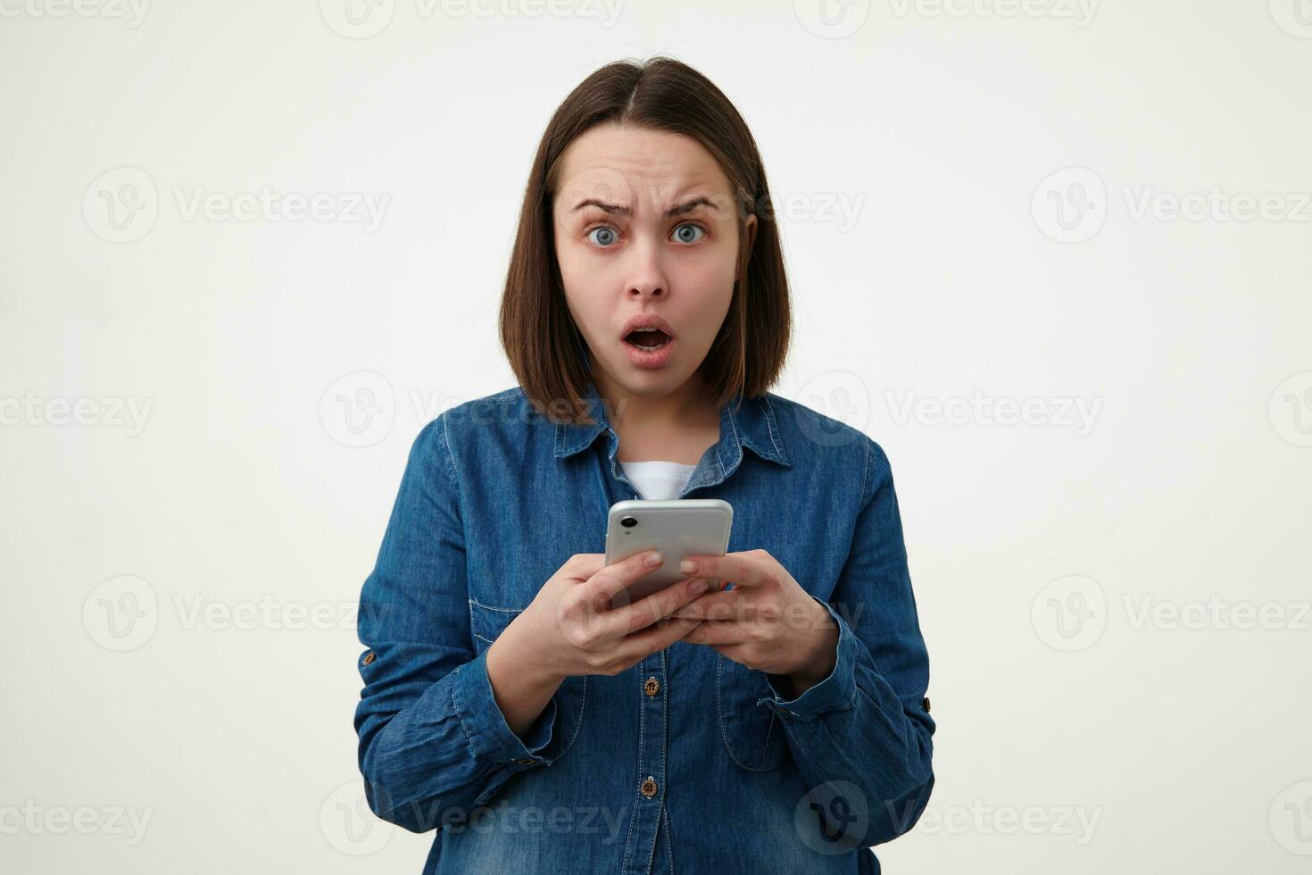 Indoor shot of shocked young dark haired lady with natural makeup looking amazedly at camera while keeping her smartphone in raised hands, posing over white background photo