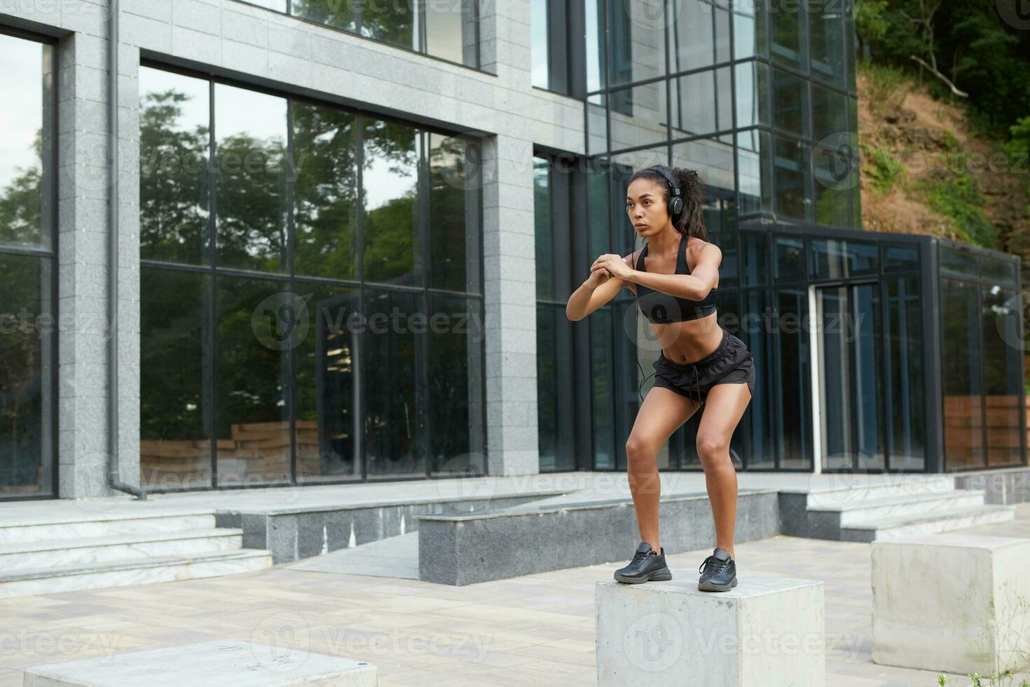 Horizontal shot of young curly dark skinned healthy woman doing evening jogging and outdoor sports exercises in city, listening to music and looking in front of herself with undistracted face photo