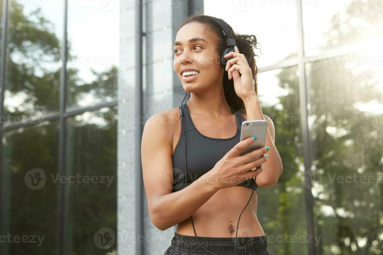 Fitness young curly model with dark skin exercising outdoors on sunny morning before working day, preparing music list for training and being in nice mood, dressed in athletic wear photo