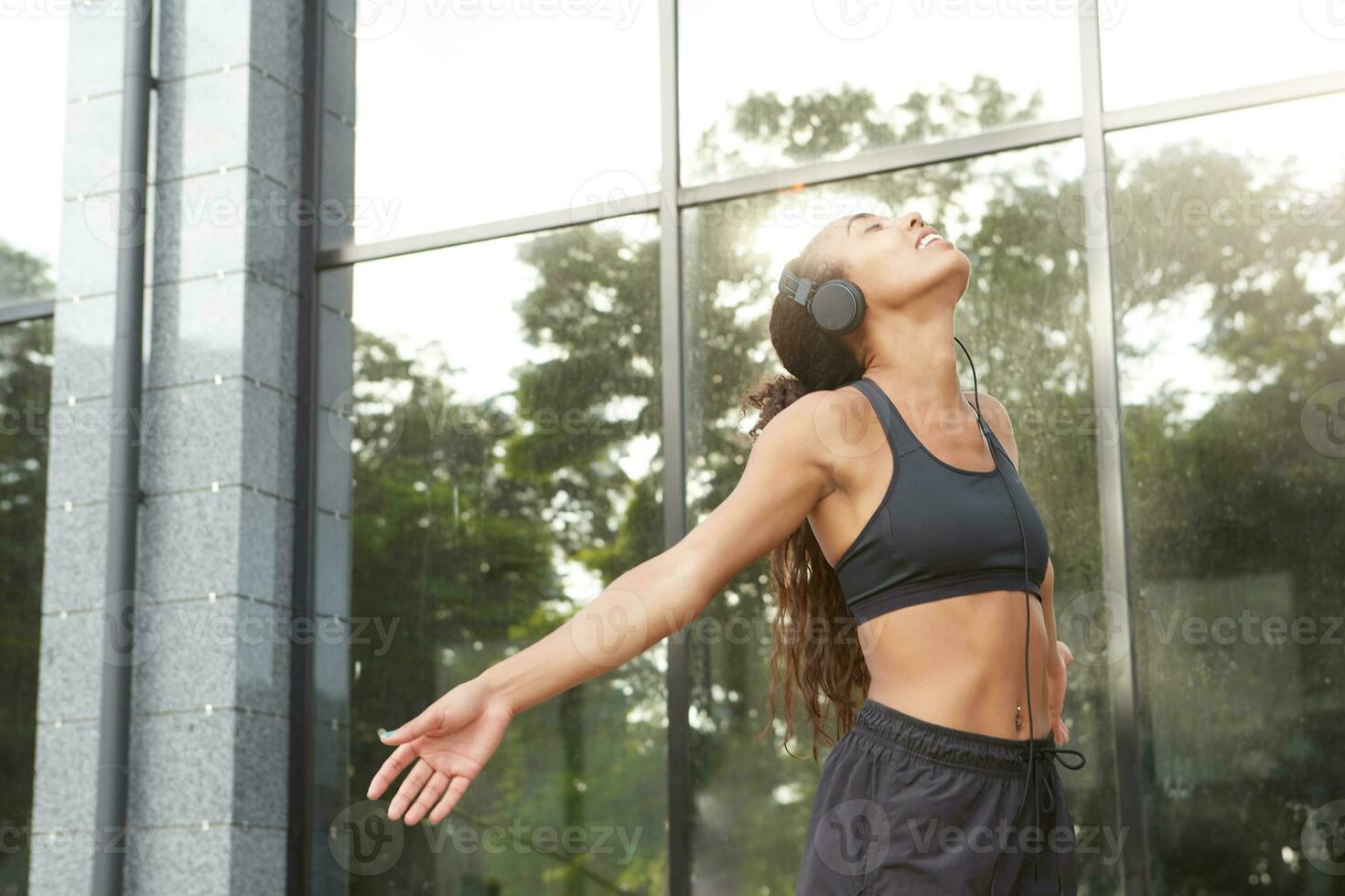 Sunny shot of cheerful healthy dark skinned female with ponytail hairstyle doing yoga fitness exercise in street, throwning back head and spreading arms, keeping eyes closed while listening to music photo