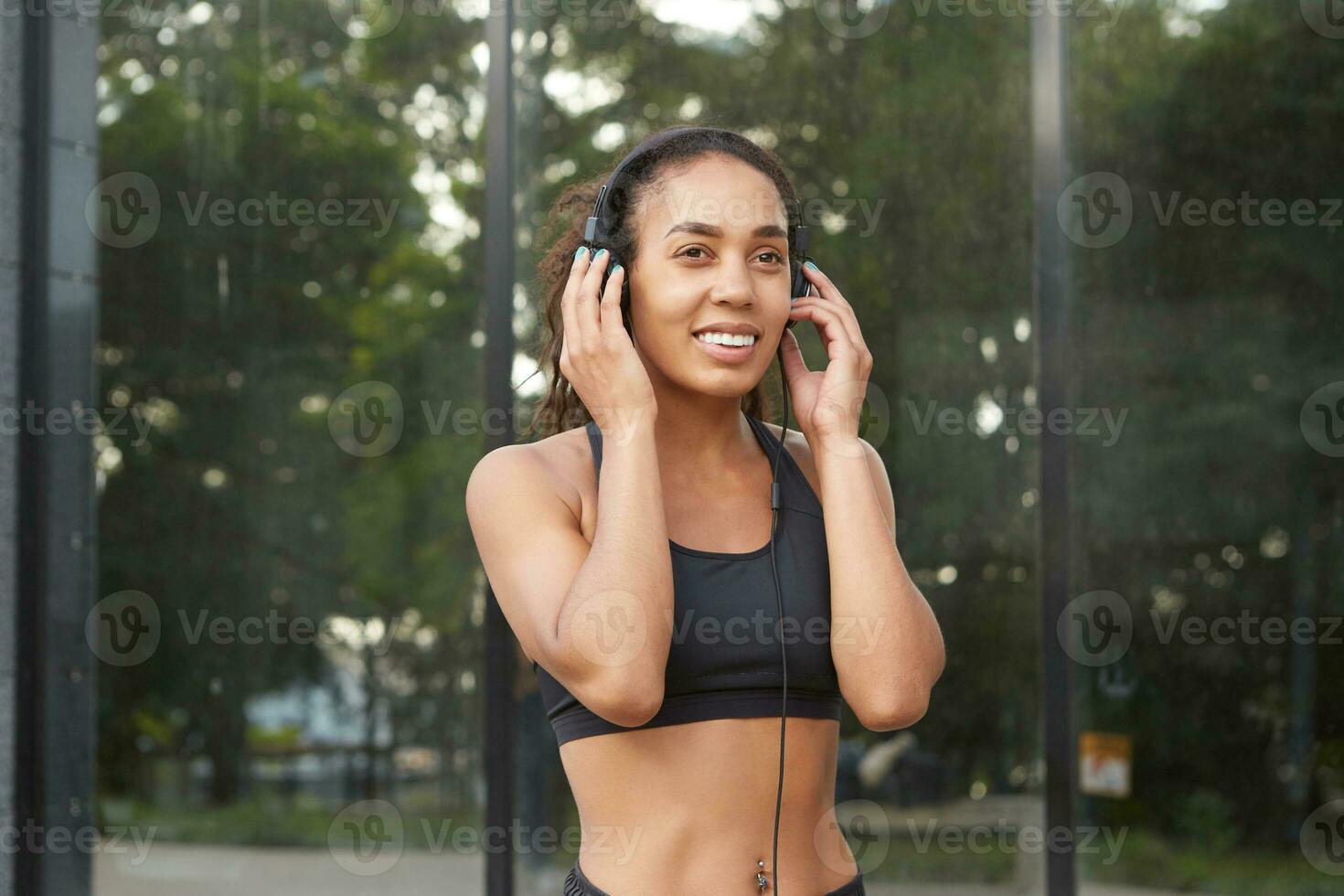 Young dark skinned curly sports woman taking break after run and enjoying favourite music, looking ahead with charming smile, goes in for sport every morning, wants to be fit and healthy photo