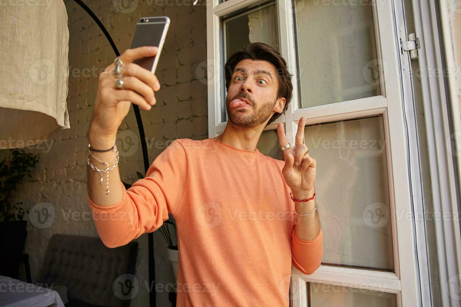 Funny indoor portrait of pretty bearded male holding phone in hand and leaning on window, making ridiculous faces and showing peace gesture, taking selfie with mobile phone photo