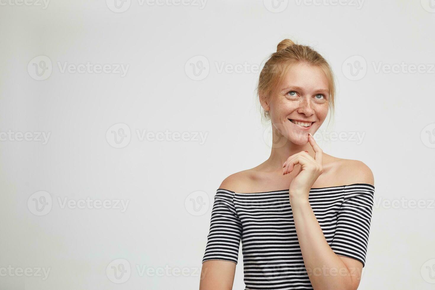Indoor photo of cheerful young pretty redhead woman biting her lips while looking cunningly upwards and holding raised index finger on her chin, posing over white background