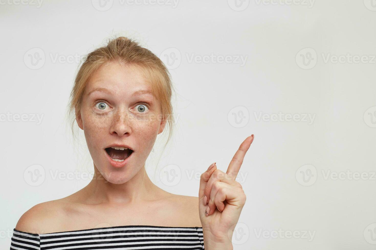 Excited young green-eyed pretty redhead female looking surprisedly at camera with wide mouth opened and raising emotionally forefinger, isolated over white background photo