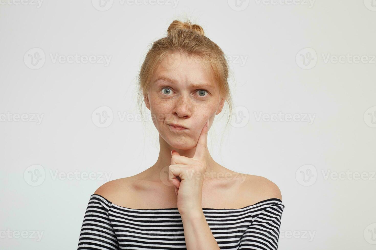 Puzzled young green-eyed redhead woman with casual hairstyle holding forefinger on her cheek and looking pensively at camera, isolated over white background photo