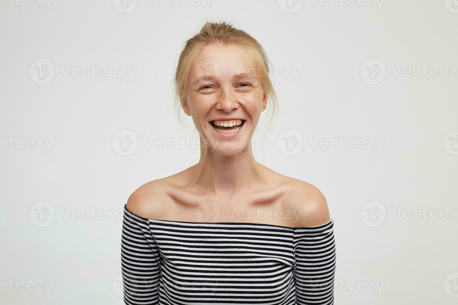 Joyful young beautiful woman wearing her foxy hair in knot while standing over white background with hands down, looking happily at camera with wide sincere smile photo