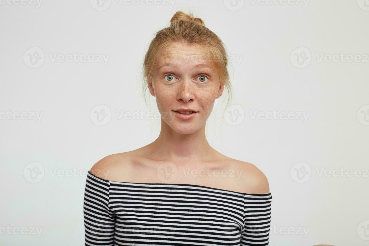 Open-eyed young lovely redhead woman with natural makeup raising surprisedly eyebrows while looking amazedly at camera, posing over white background with hands down photo