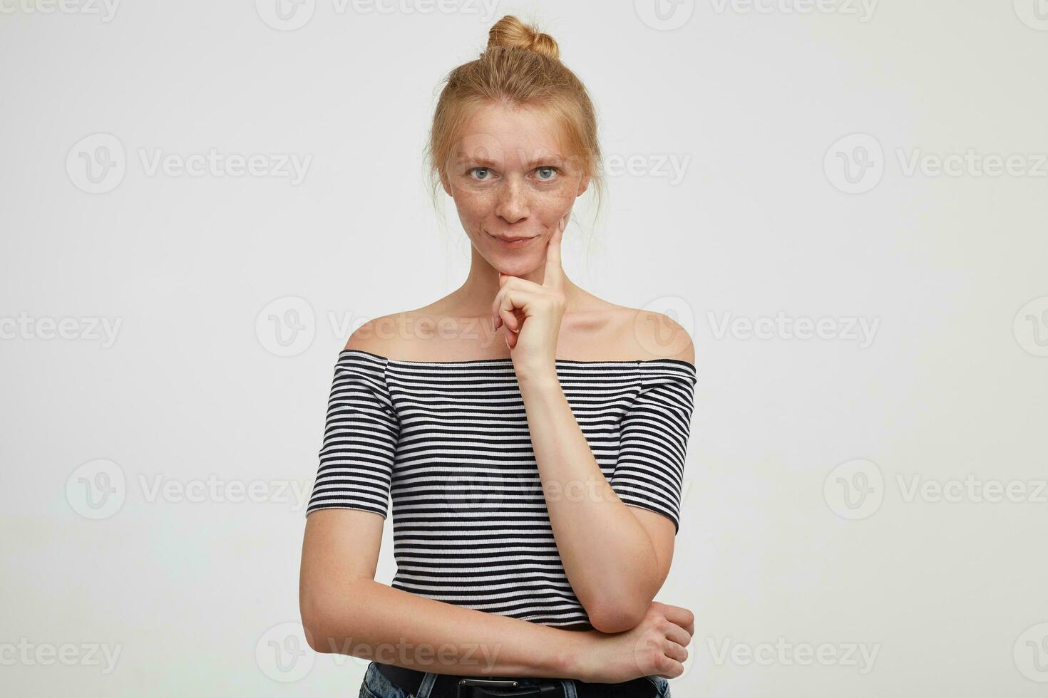 Pensive young green-eyed redhead female with casual hairstyle keeping her lips folded while looking at camera and holding forefinger on her cheek, posing over white background photo